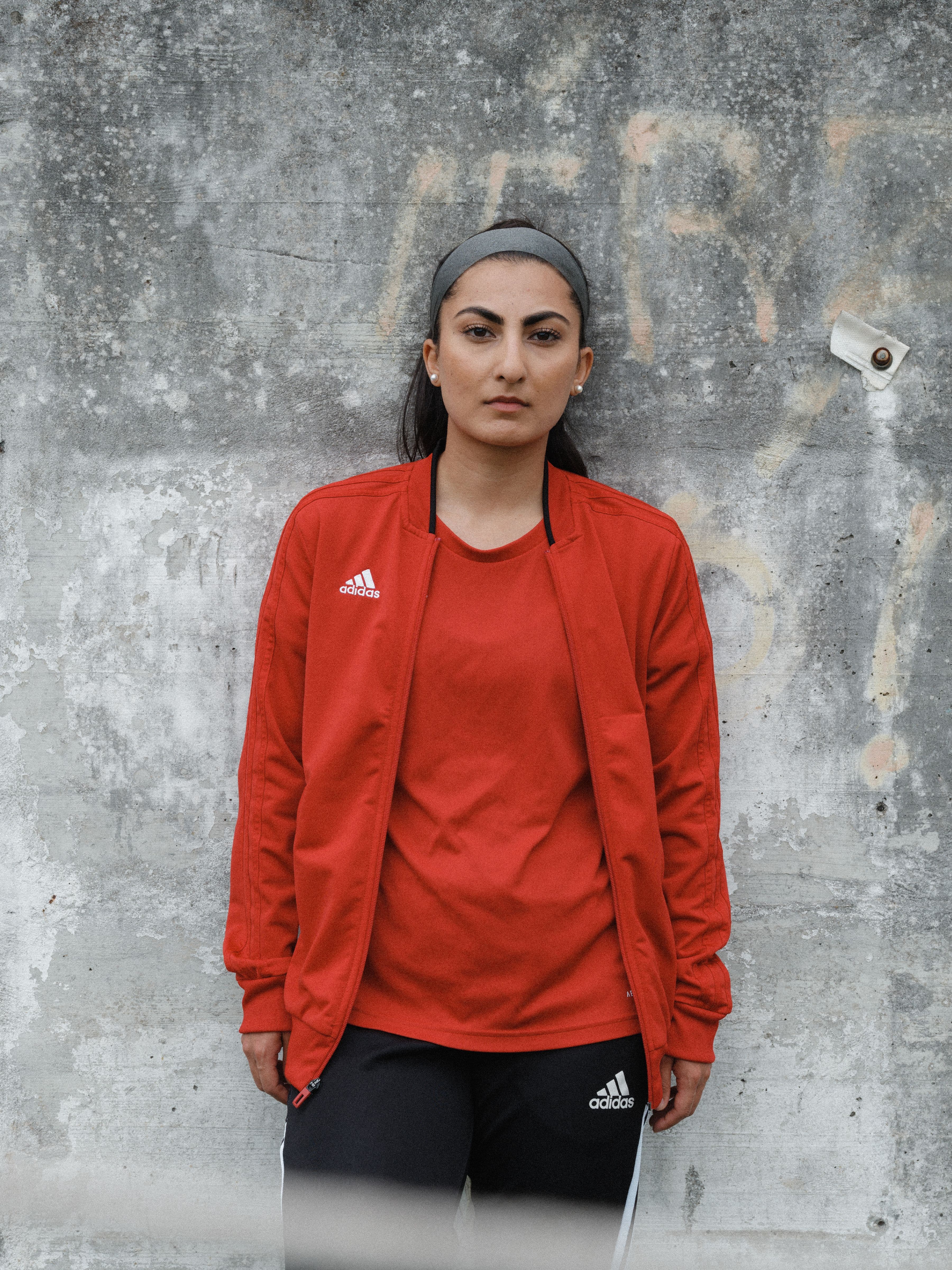 Person in a red Adidas jacket and black pants stands against a weathered concrete wall, looking directly at the camera.