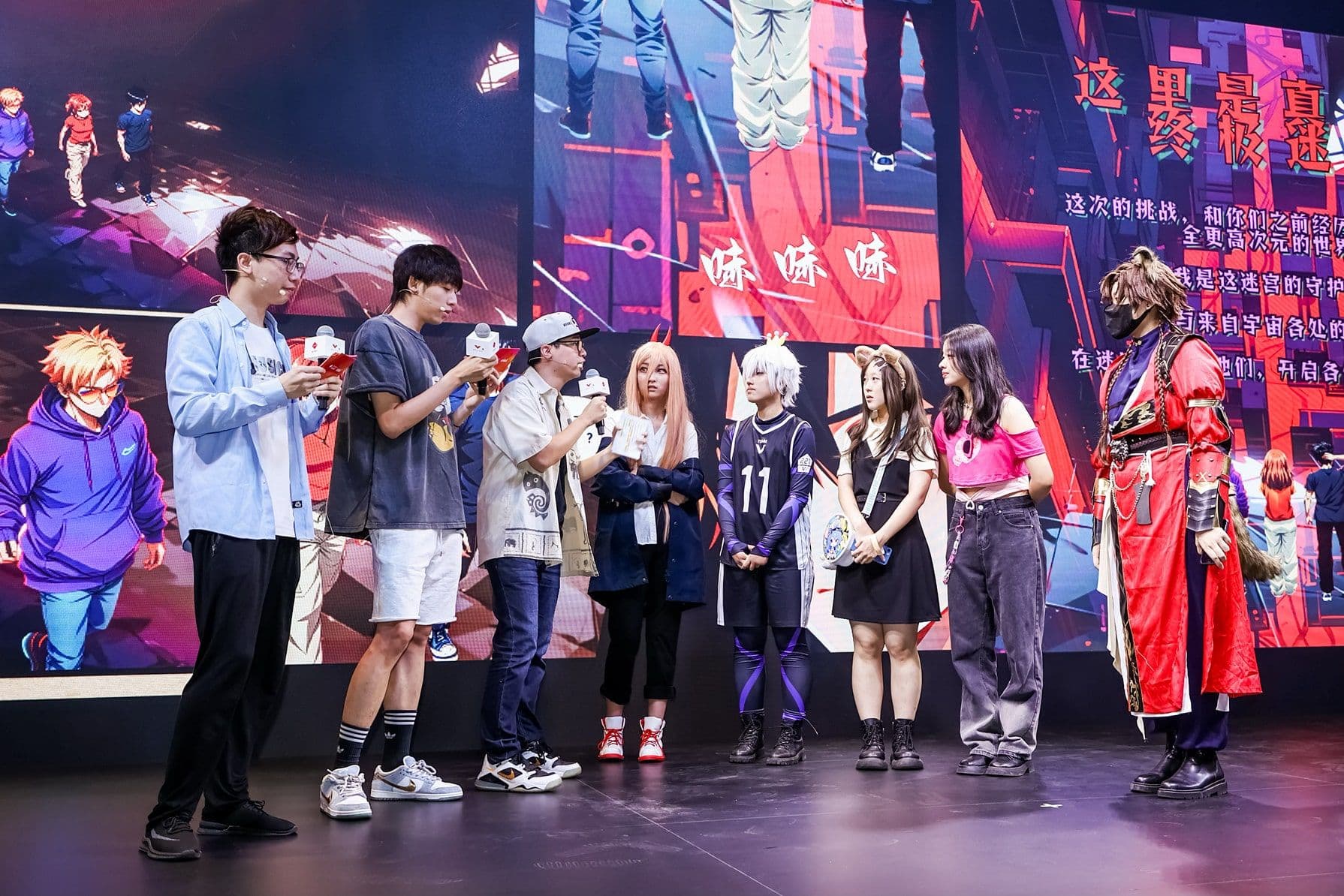 A group of people on stage, some in cosplay, stand in front of a large, colorful anime-themed screen, engaging with the audience.