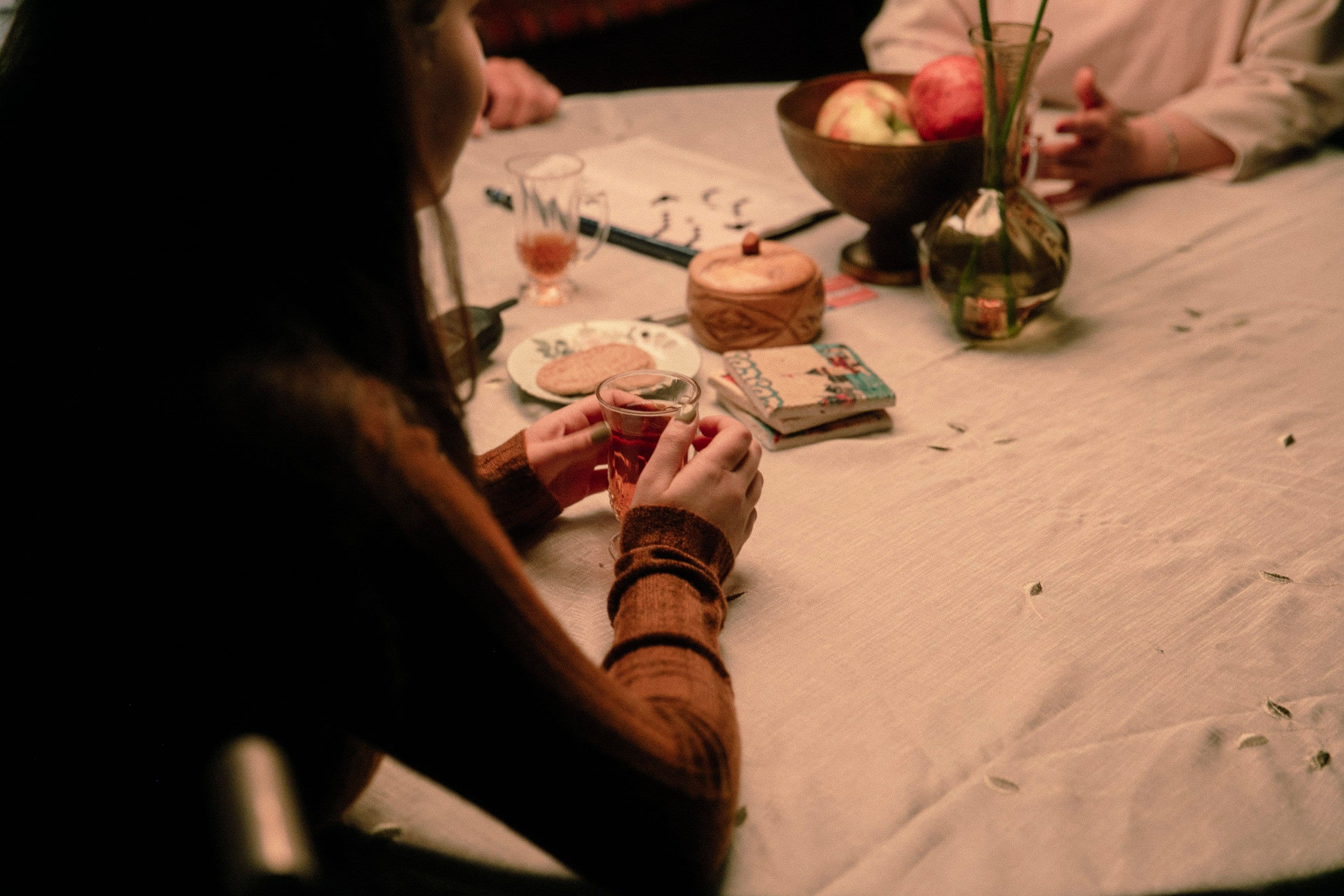 Close up of two people sitting at a table with a white cloth, holding drinks. A bowl of apples, a vase, and coasters are on the table. Warm lighting.