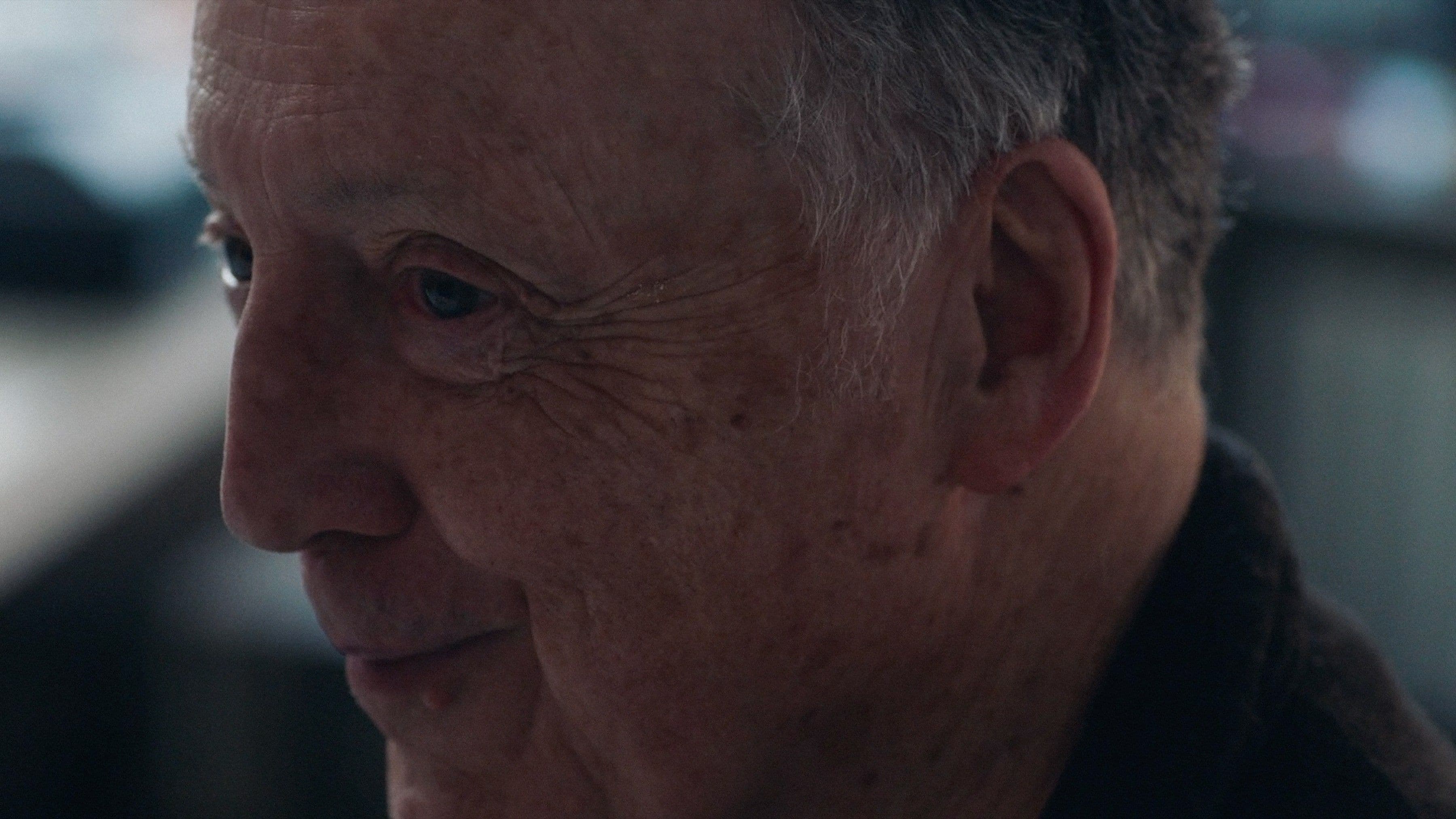 Close up of man with gray hair, smiling slightly, in a dimly lit indoor setting with blurred background.