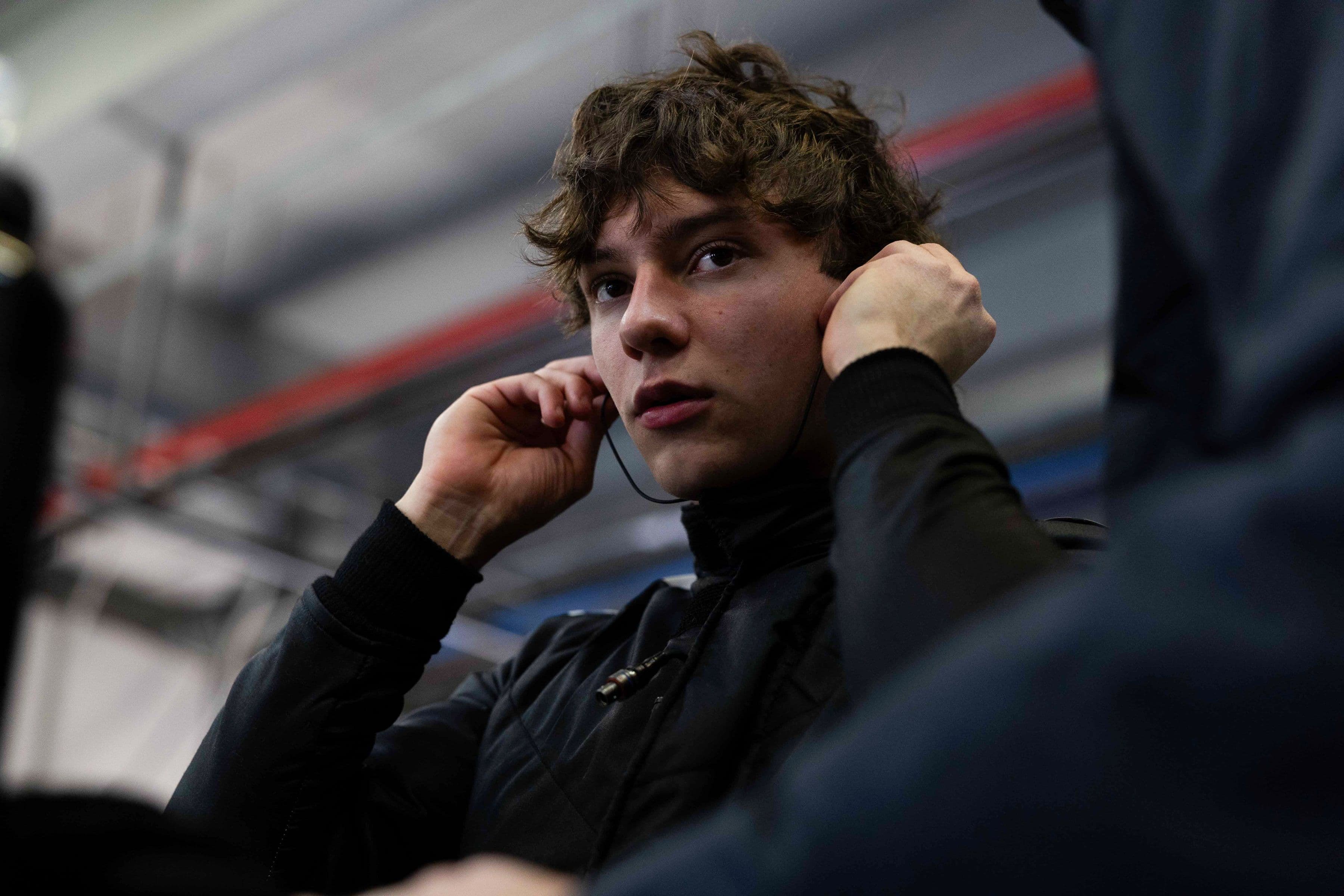 Young man with curly hair adjusts earbuds, wearing a black jacket in a car garage.