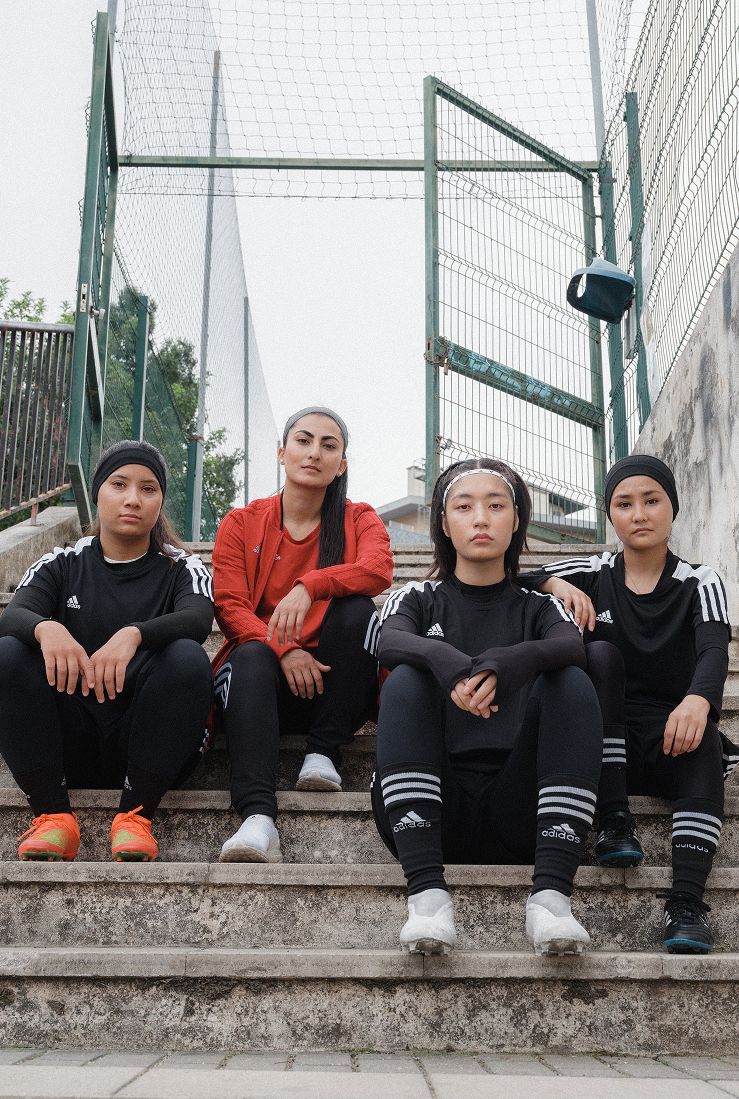 Four women in sportswear sit on outdoor steps. Three wear black outfits, one in a red jacket. Fenced court in background.