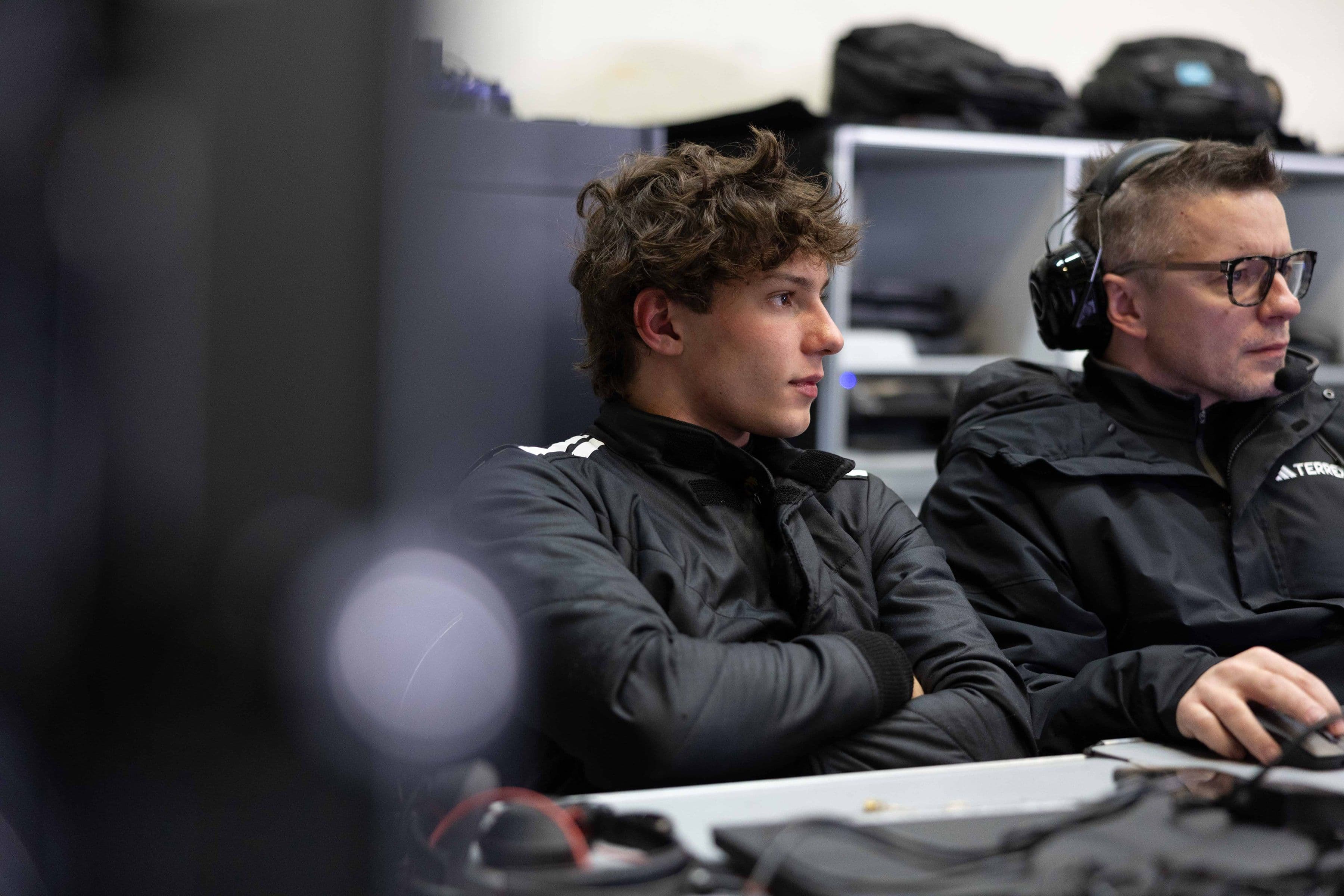 Two men in black jackets sit at a desk with computers in a car garage, focused on test results. One wears headphones. Indoor setting.