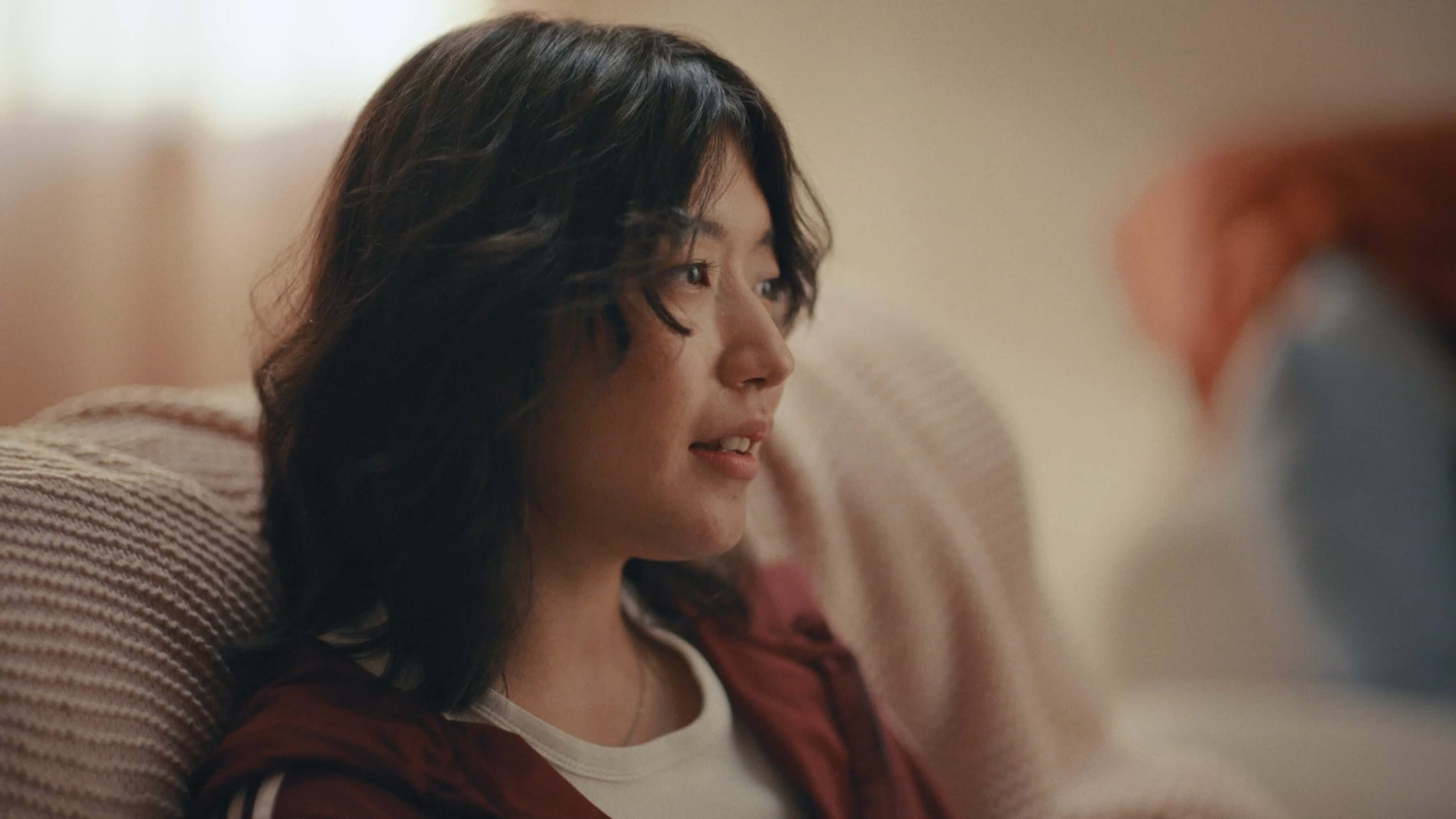 Woman with long dark hair, wearing a maroon top, sits on a beige couch, looking to the side in a softly lit room.