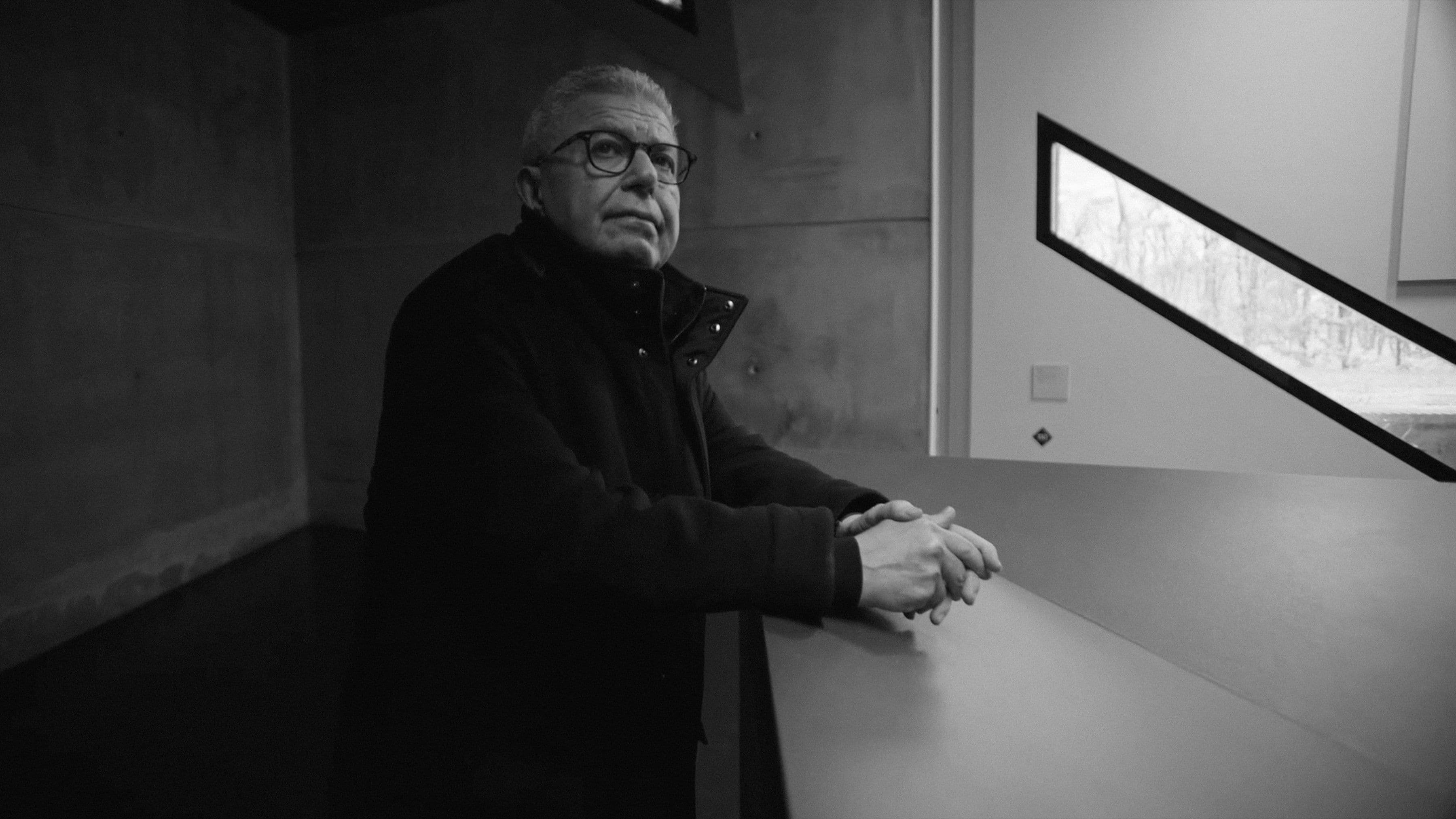 Man in glasses and coat stands by a modern, angular table.
