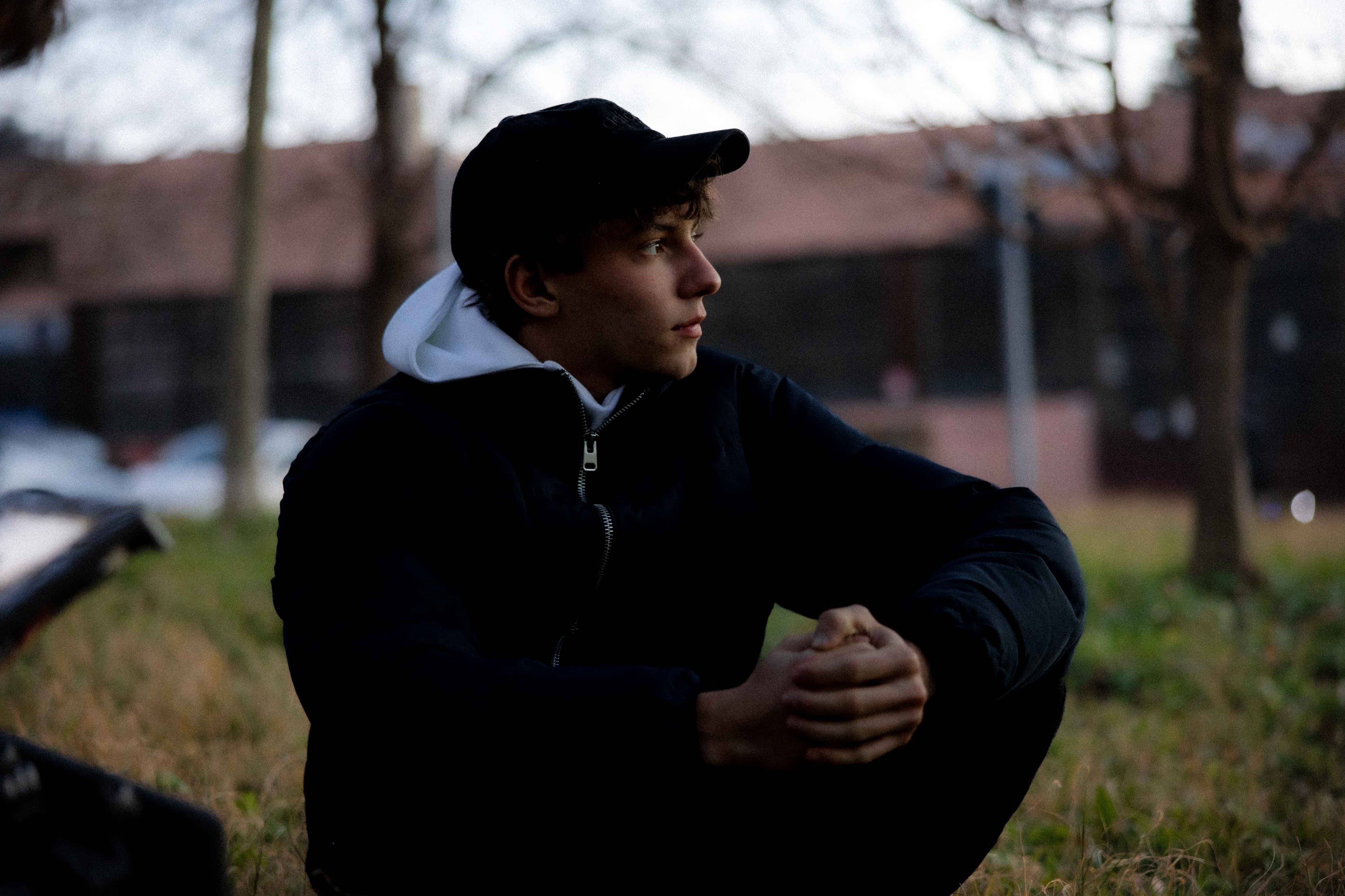 Young man in black jacket and cap sits on grass, hands clasped, looking contemplative. Overcast sky, bare trees in background.