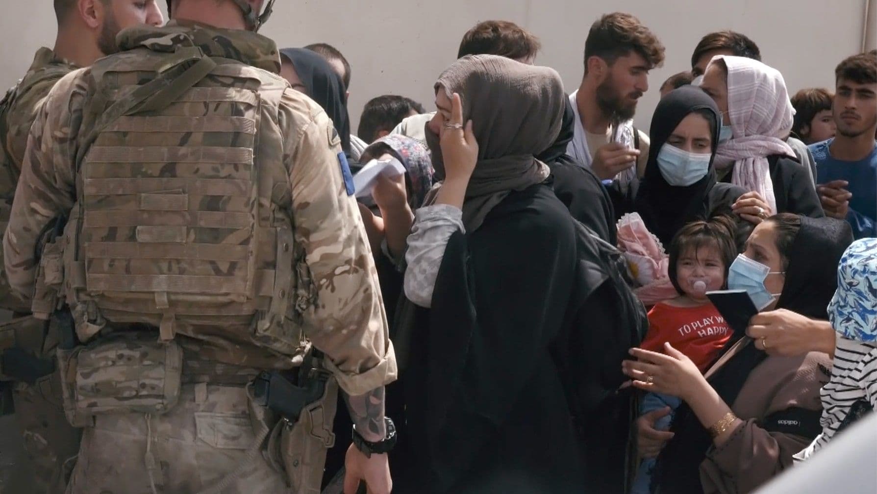 A soldier in camouflage gear faces a crowd of people, including women and children in hijabs and masks.