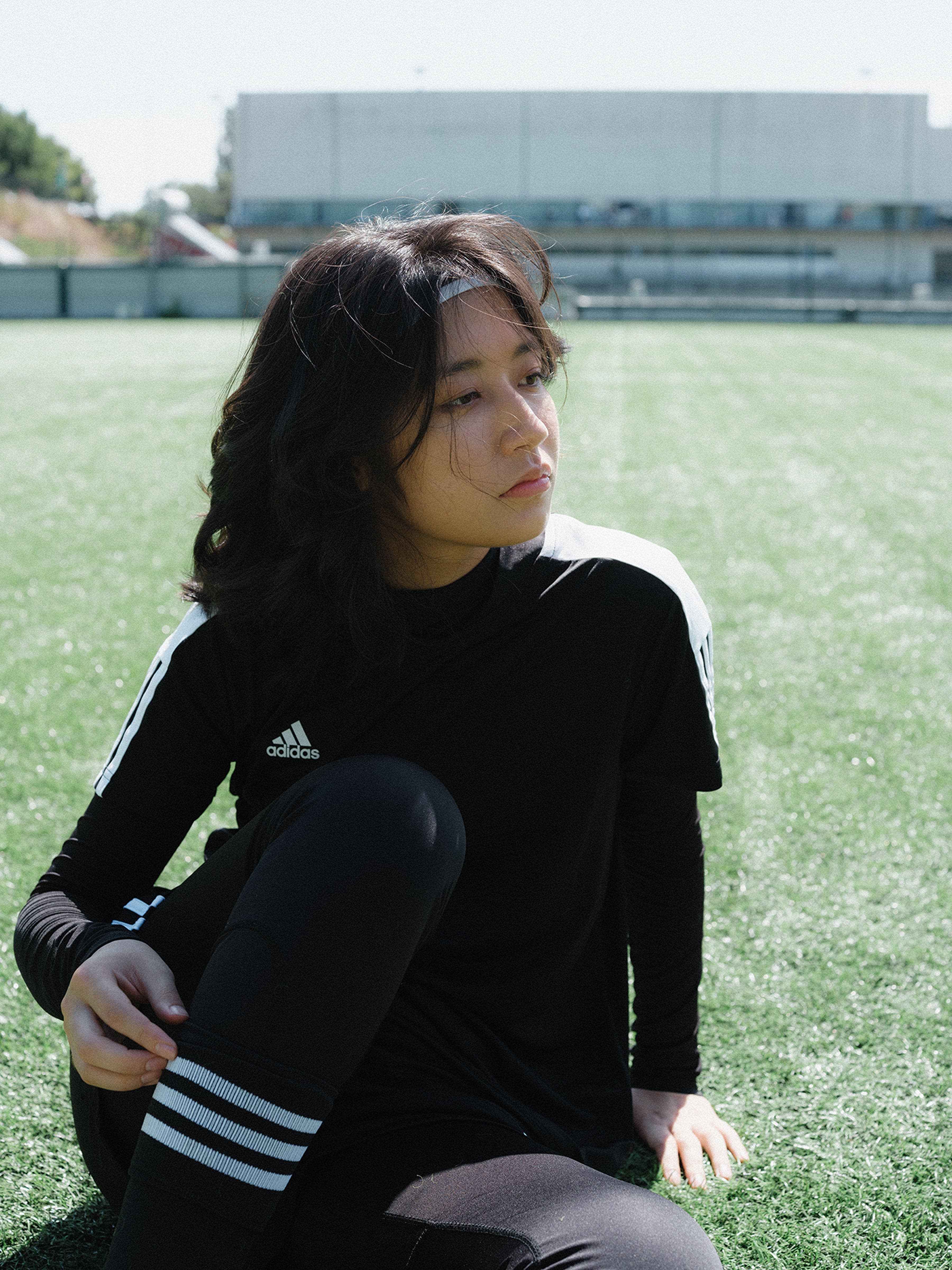 Young woman in black Adidas sportswear sits on green soccer field, looking thoughtful. Bright, sunny day with a building in background.