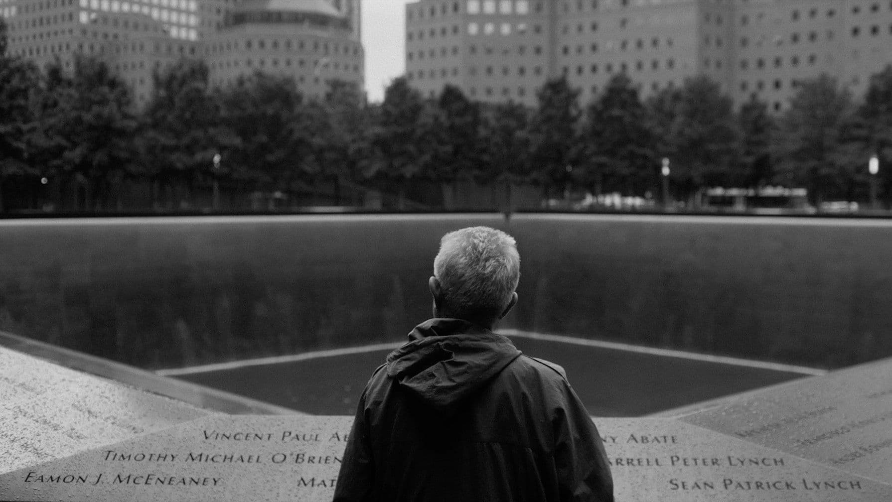 A person with short gray hair stands at a memorial pool, surrounded by engraved names, in an urban setting with trees and buildings.