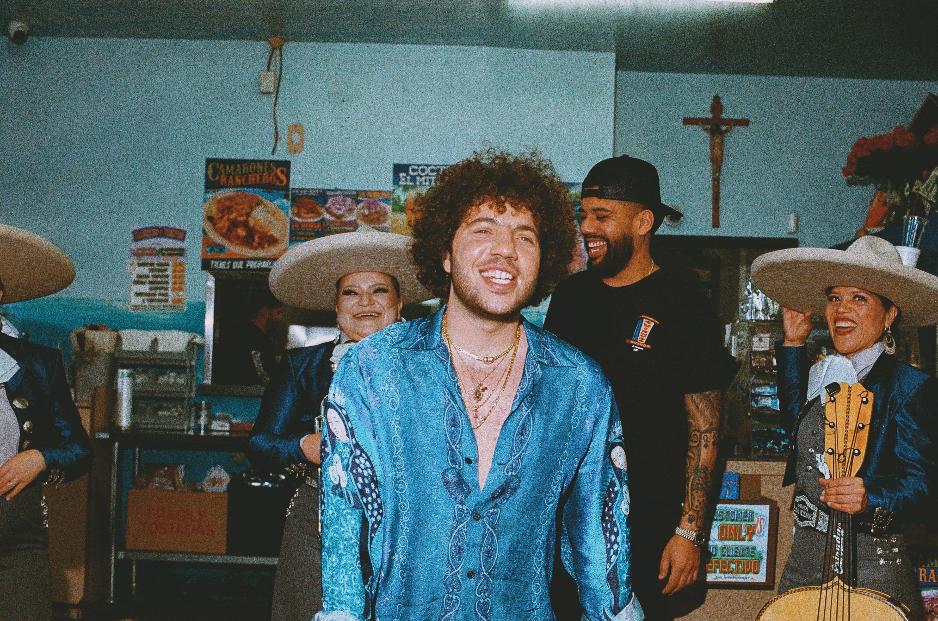 A man in a blue patterned shirt smiles in a lively restaurant with mariachi musicians in sombreros and a man in a black cap.