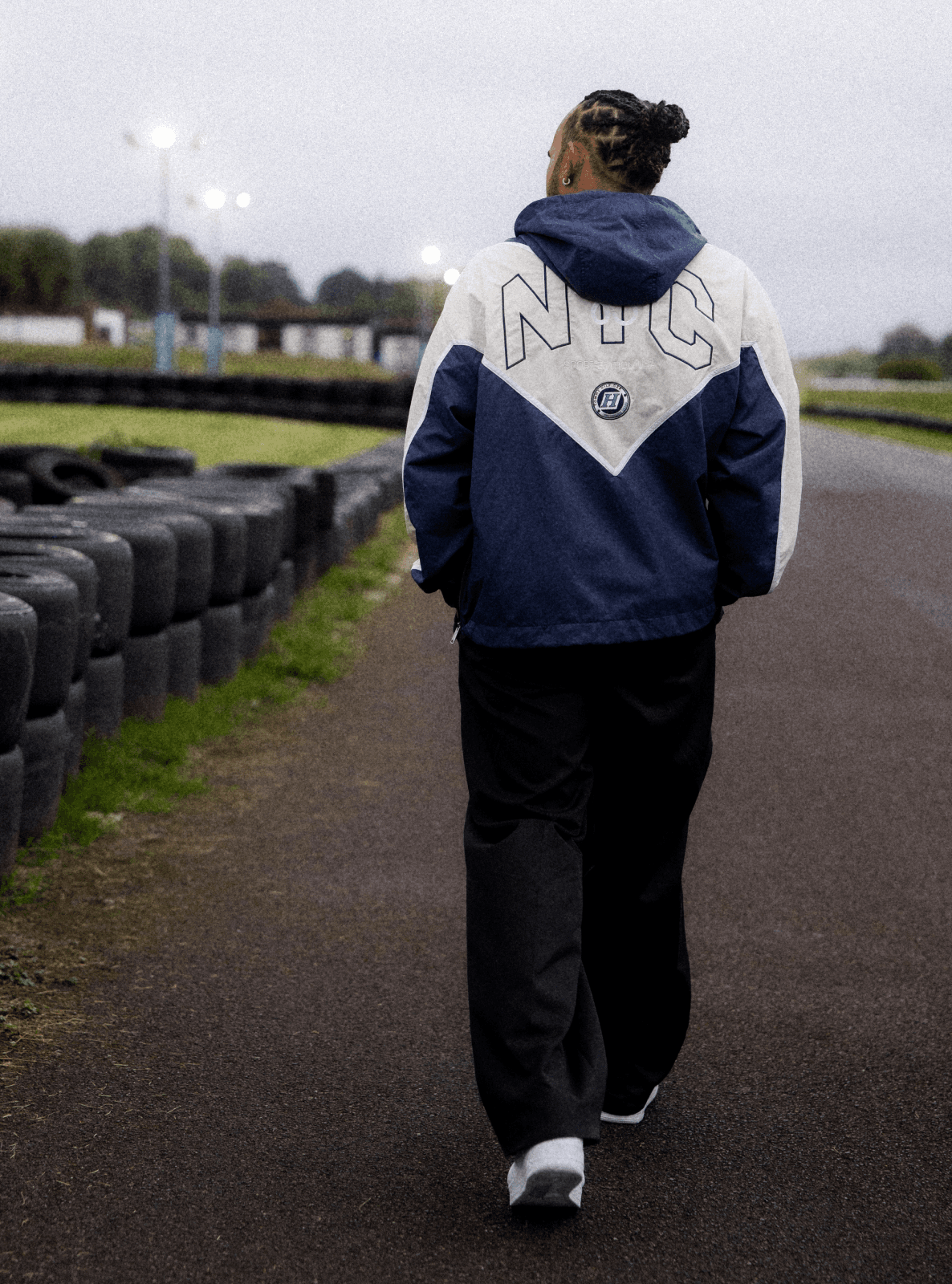 Person with braided hair walking on an karting track, wearing a blue and white "NYC" jacket, near stacked tires and grassy area.