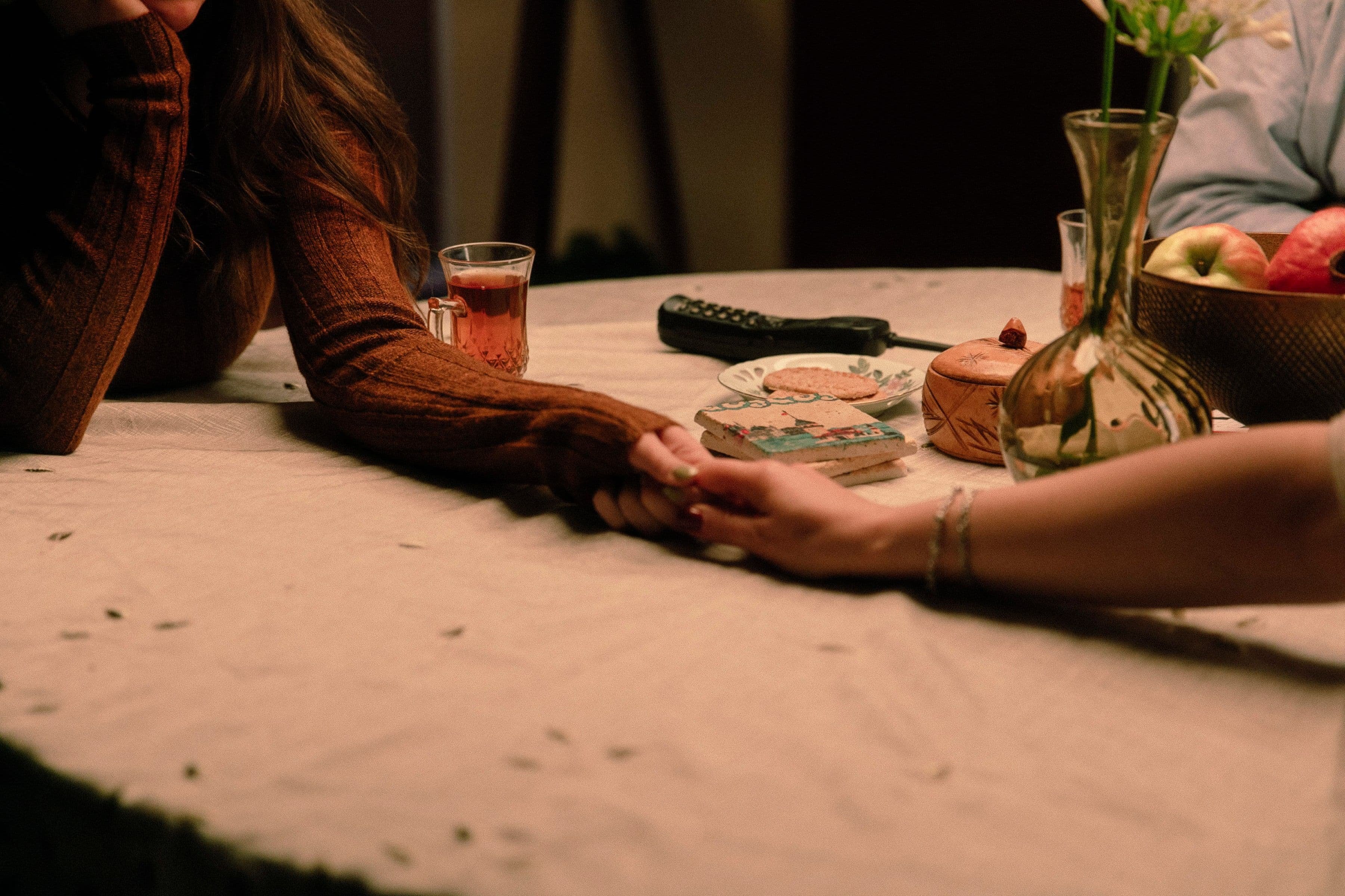 Close up of two people holding hands across a table with a beige cloth, surrounded by a vase, fruit bowl, remote, and tea glass.