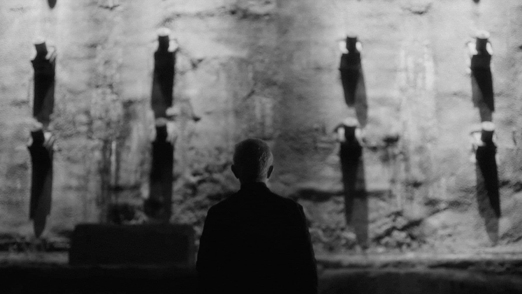 Silhouette of a person facing a textured stone wall with vertical shadows, in a dimly lit, monochrome setting.