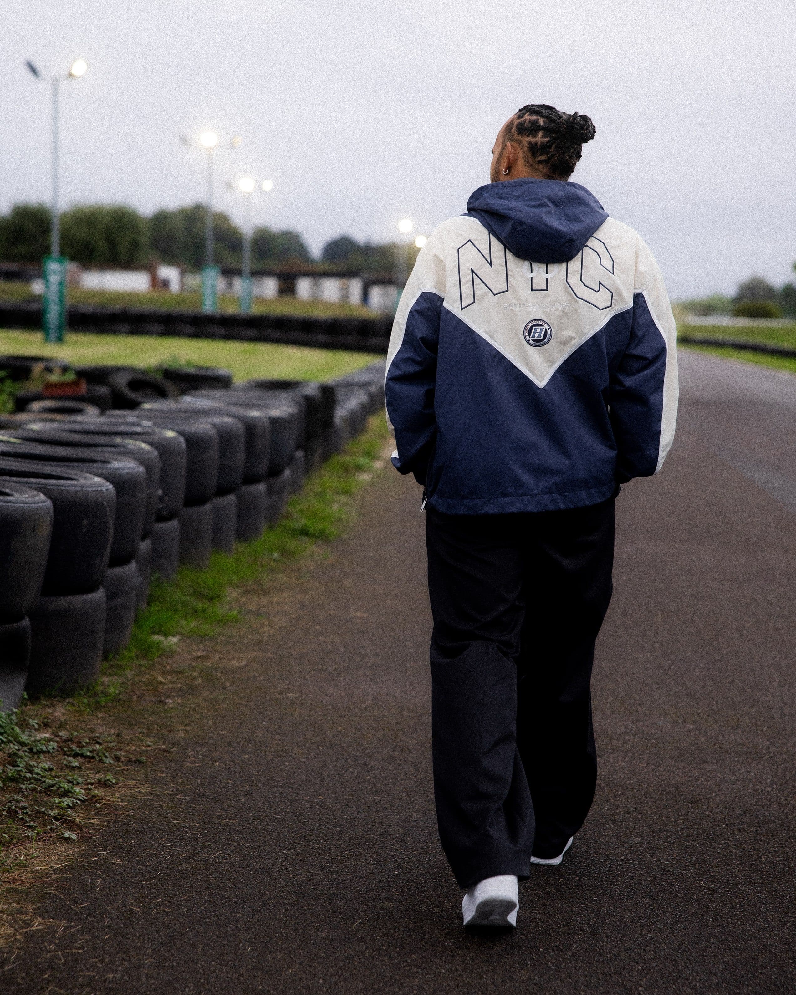 Person with braided hair walking on an karting track, wearing a blue and white "NYC" jacket, near stacked tires and grassy area.