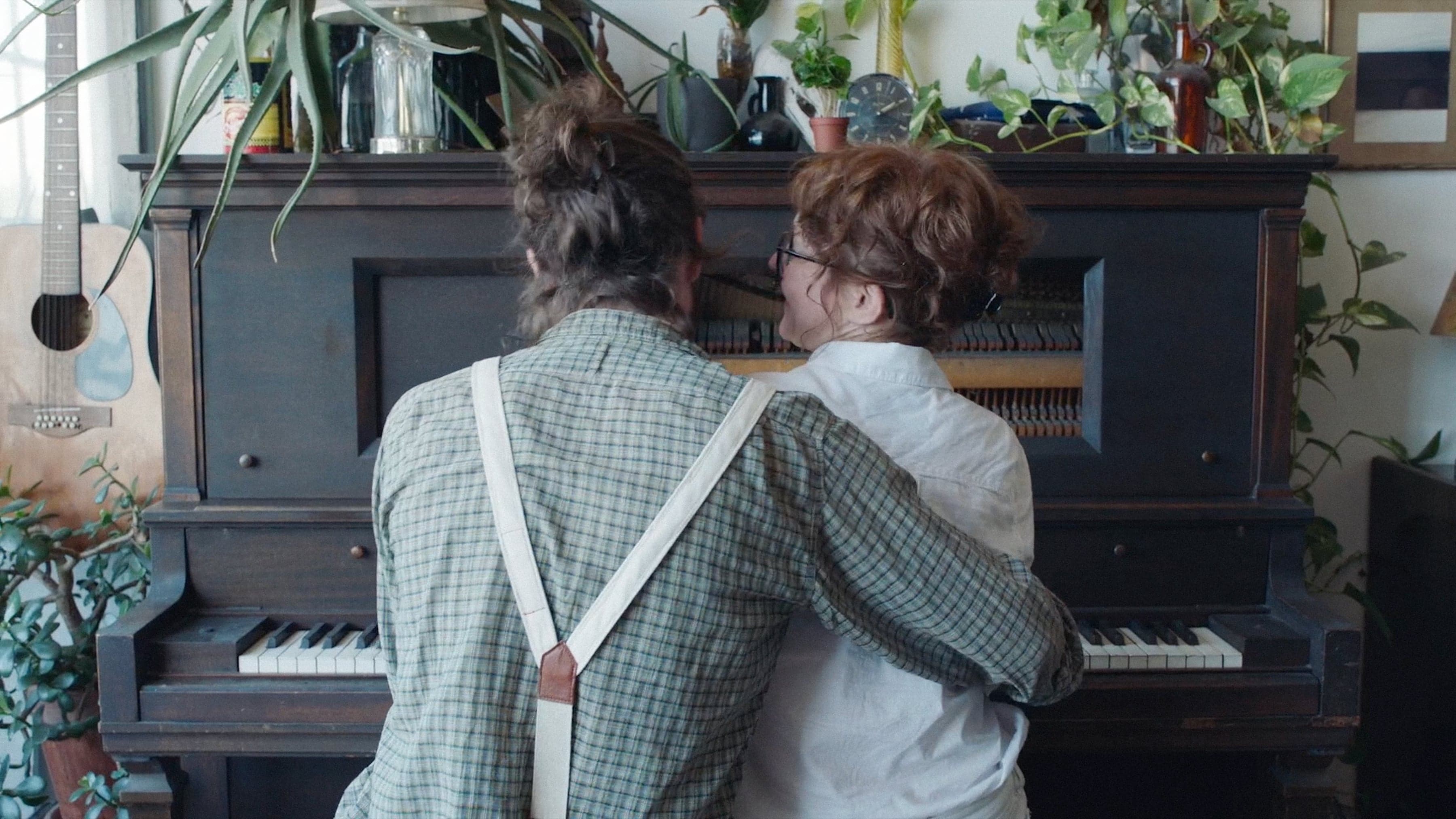 Two people with their backs to the camera sit at a piano, surrounded by green plants. One wears suspenders, the other glasses.