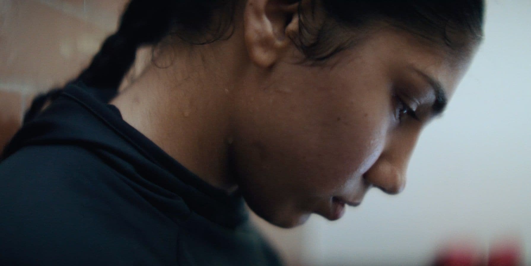 Close-up of a woman with braided hair, wearing a dark hoodie, leaning against a brick wall, focused and slightly sweaty.