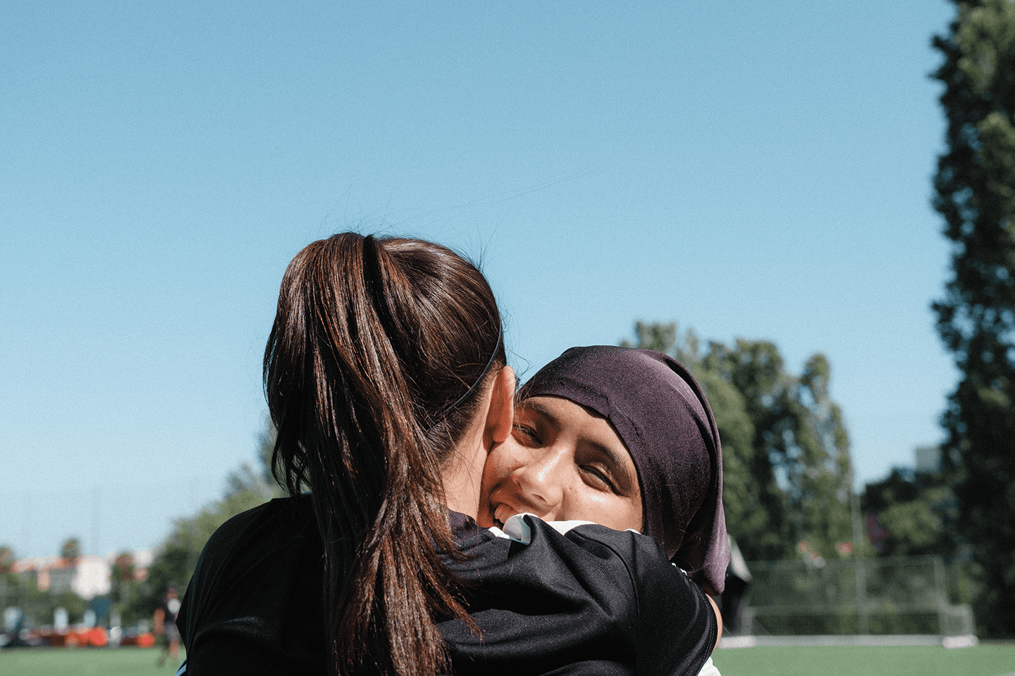 Two women in sports attire embrace on a sunny soccer field. One wears a black hijab. Trees and clear blue sky in the background.
