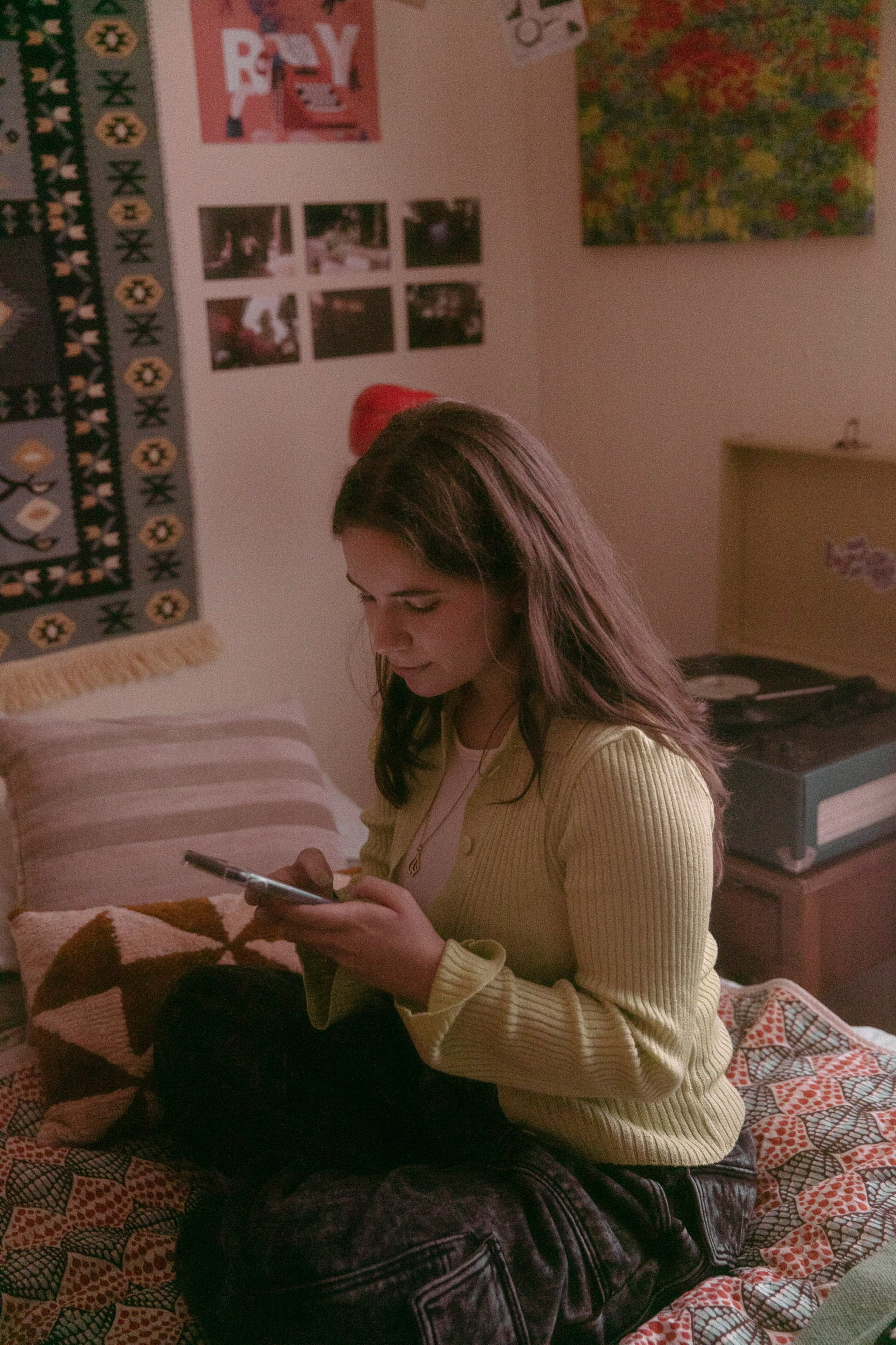Young woman in a yellow sweater sits on a bed, using a cell phone. Room has colorful wall art and a vintage record player.