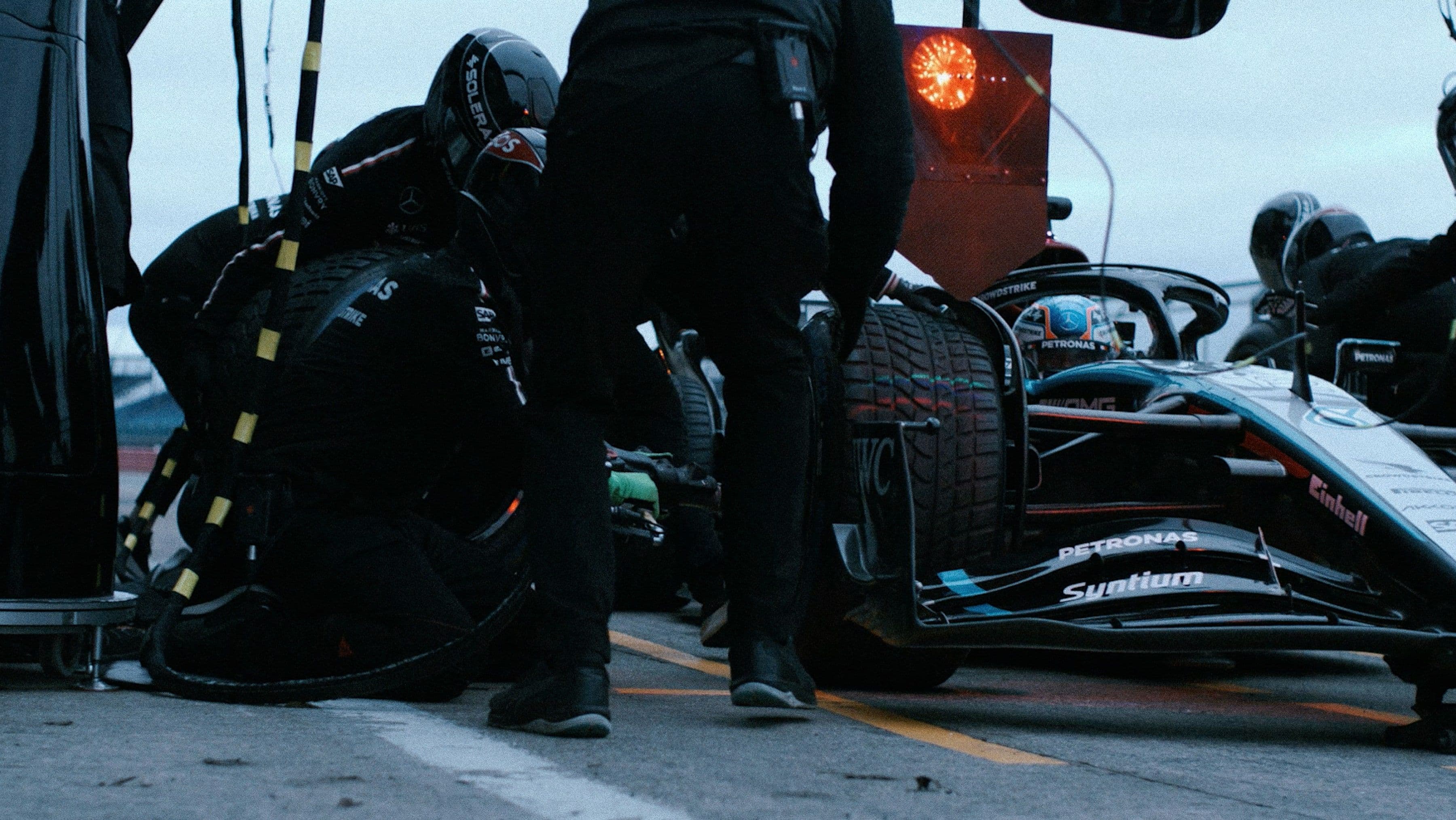 Race car pit stop with crew in black suits changing tires. Car is white with blue accents.