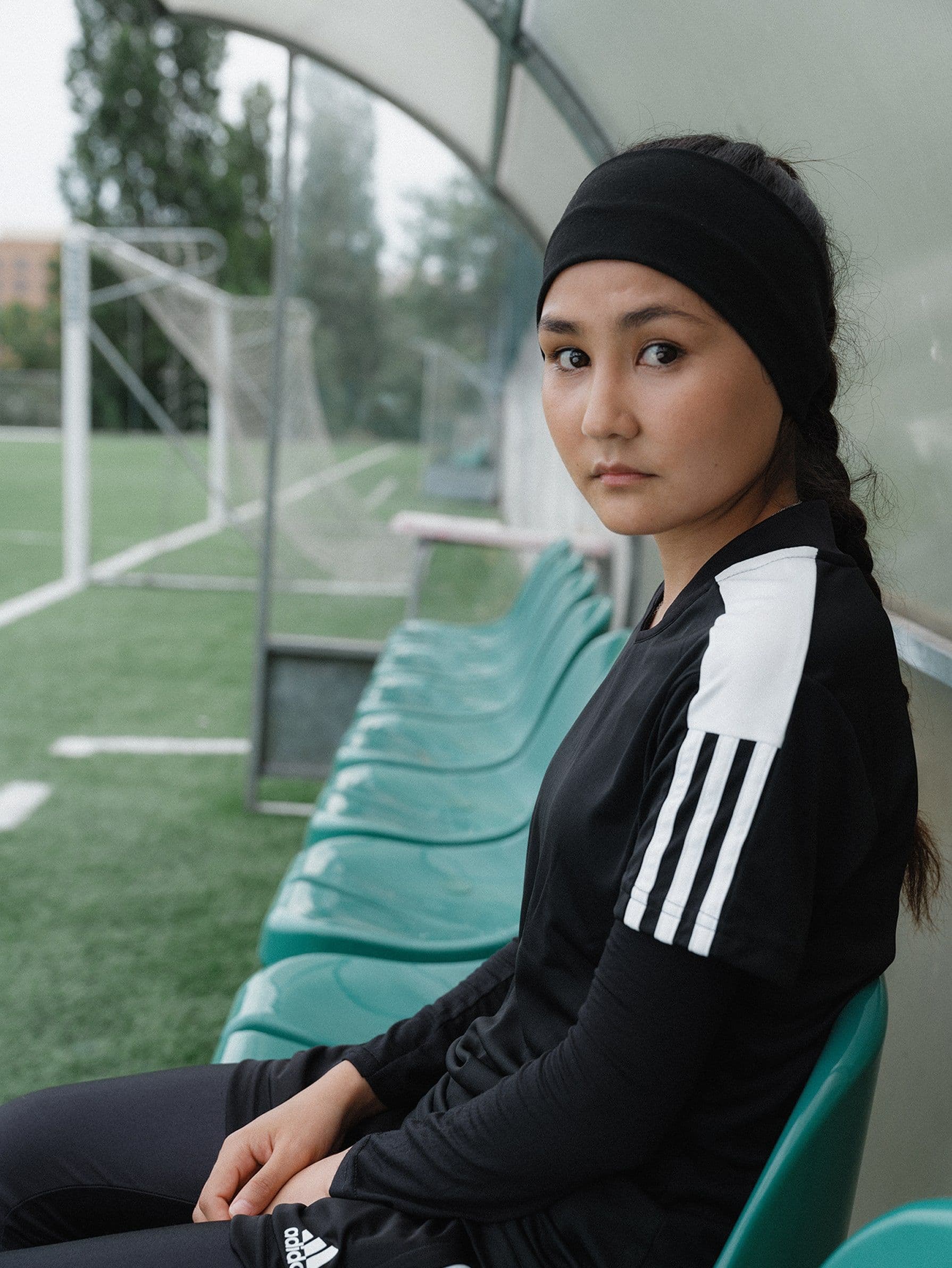 Young woman in black sportswear and headband sits on green bench in soccer field, looking towards the camera.