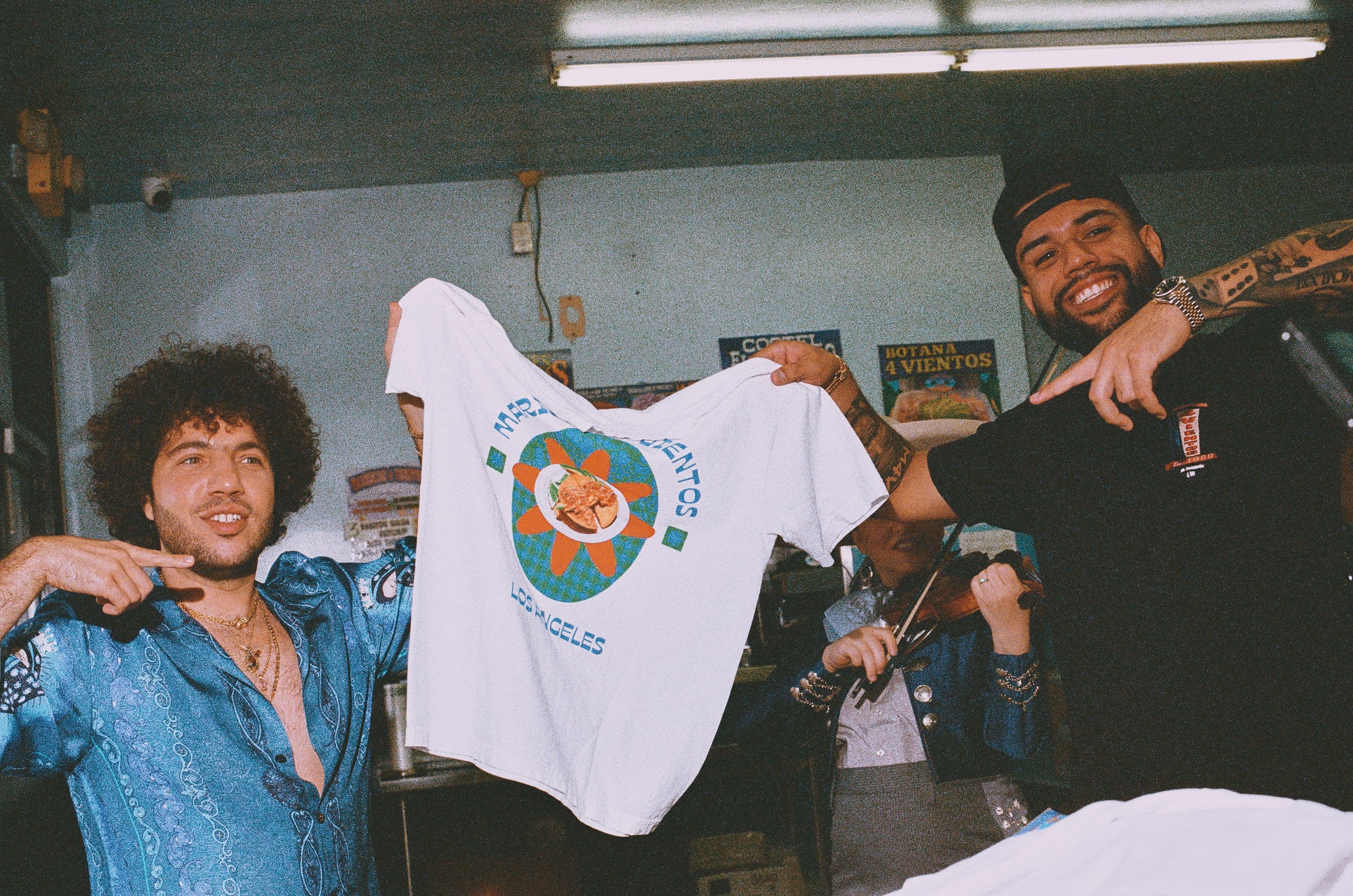 Two men in a restaurant hold up a white T-shirt with colorful graphics. One wears a blue shirt, the other a black cap and shirt.