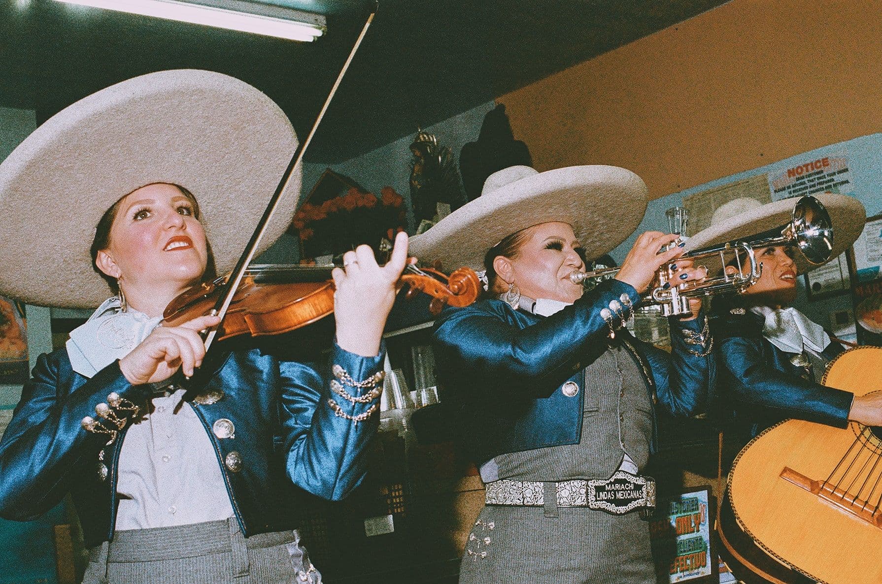 Three women in blue mariachi outfits play violin, trumpet, and guitar indoors. They wear large beige sombreros, performing energetically.