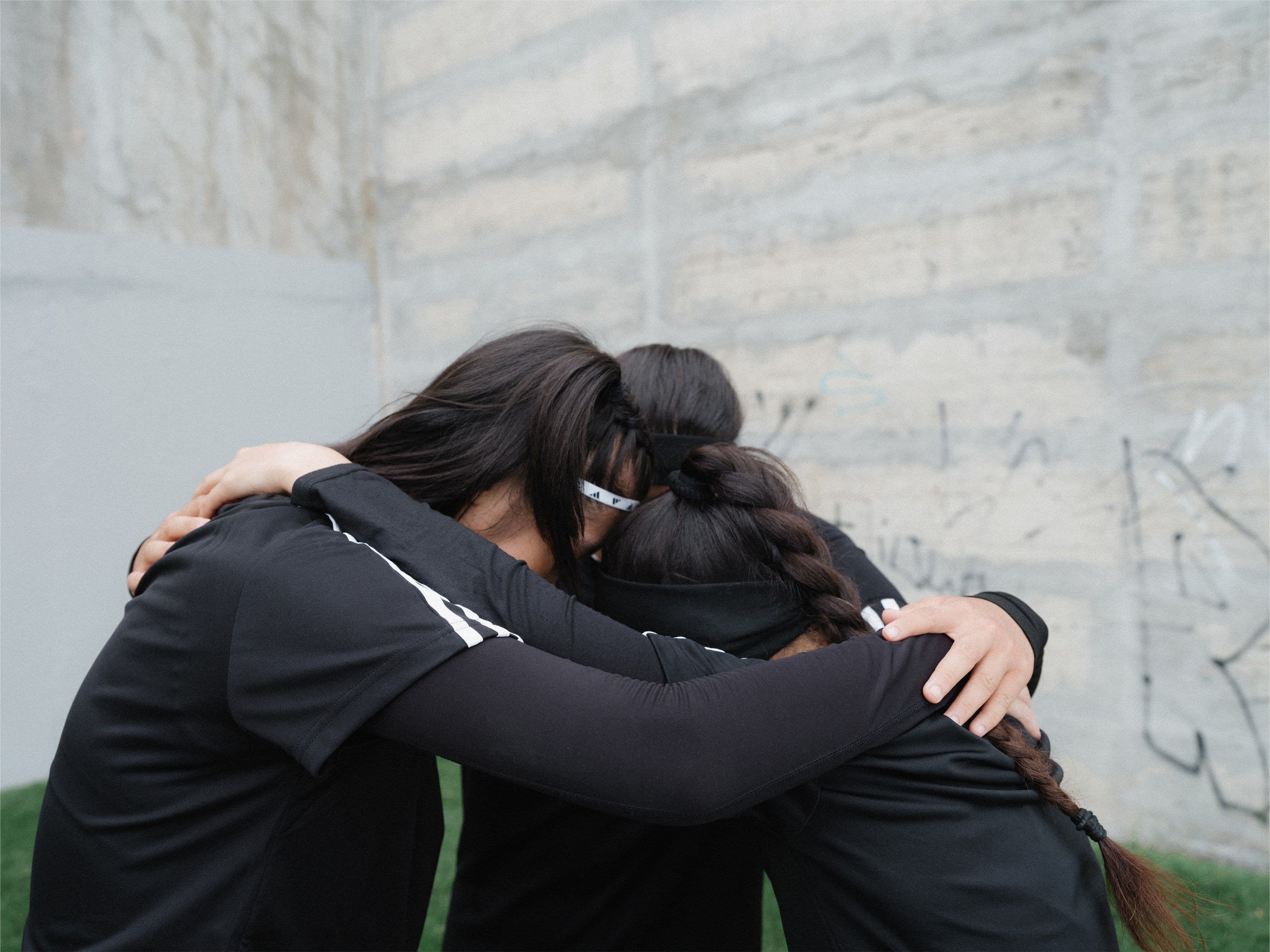 Three people in black athletic wear huddle closely, arms around each other, against a concrete wall with graffiti.