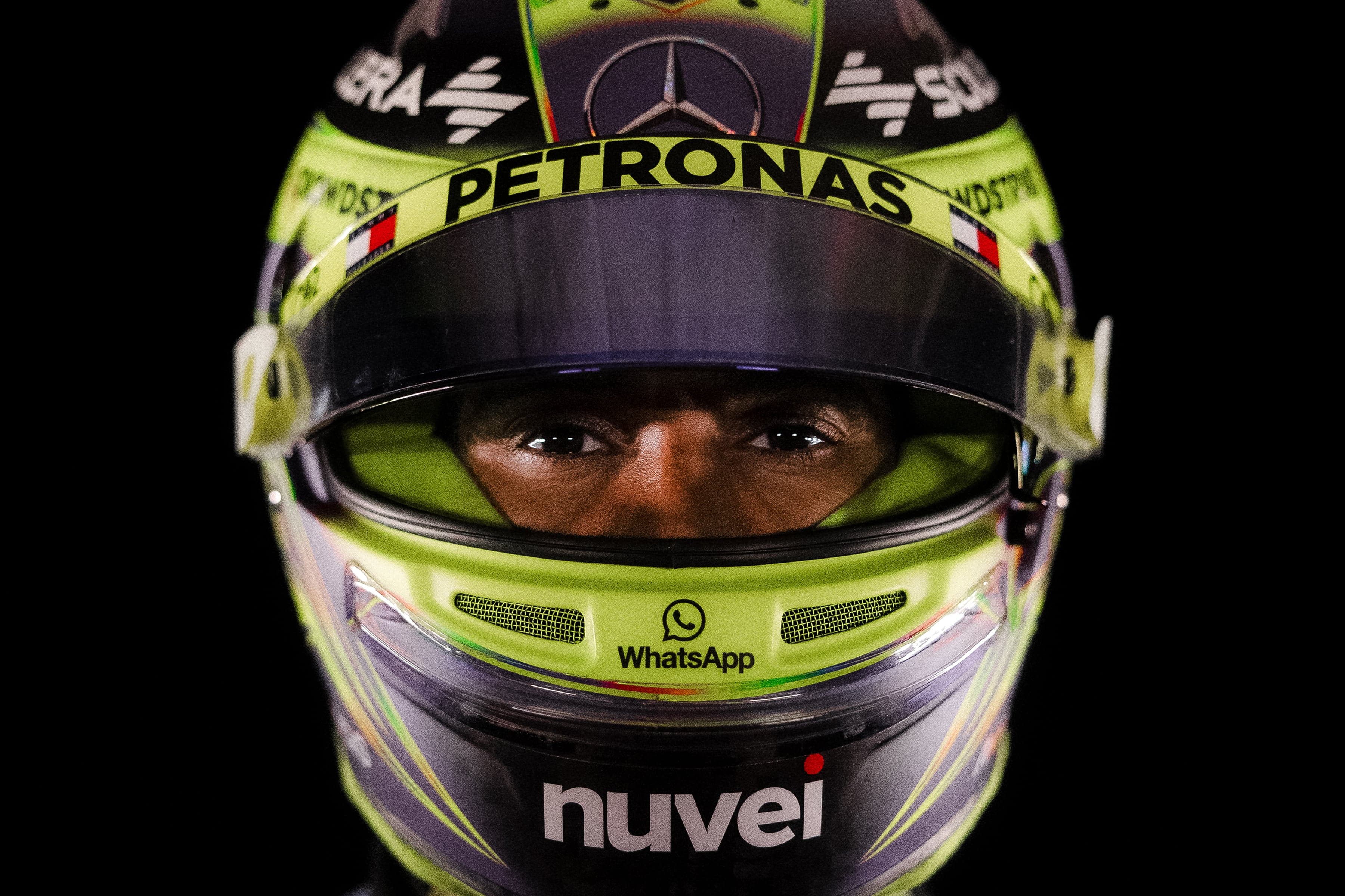 F1 driver wearing a neon green Mercedes helmet with "Petronas" and "WhatsApp" logos, focused eyes visible, dark background.