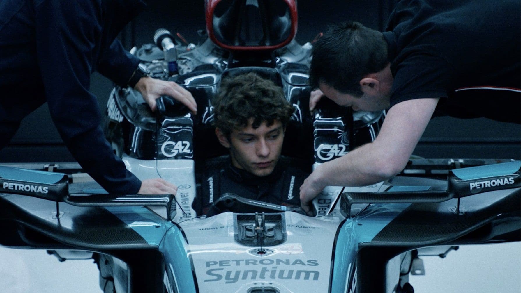 Young man sits inside an F1 car, surrounded by two technicians adjusting controls.