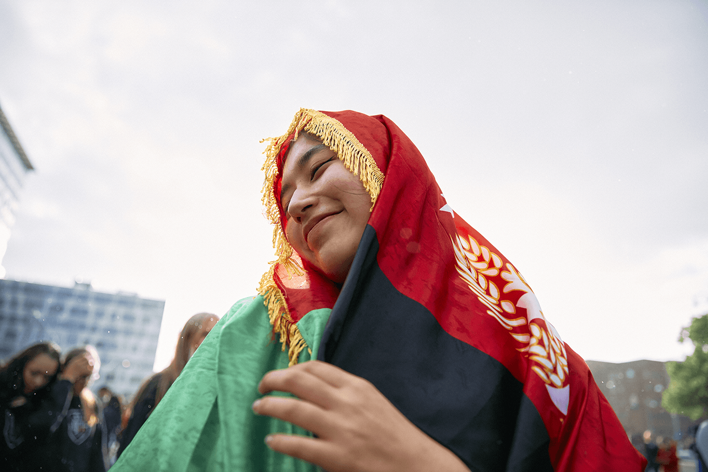 Smiling person wrapped in a red, black, and green flag with gold fringe, standing outdoors under a cloudy sky.