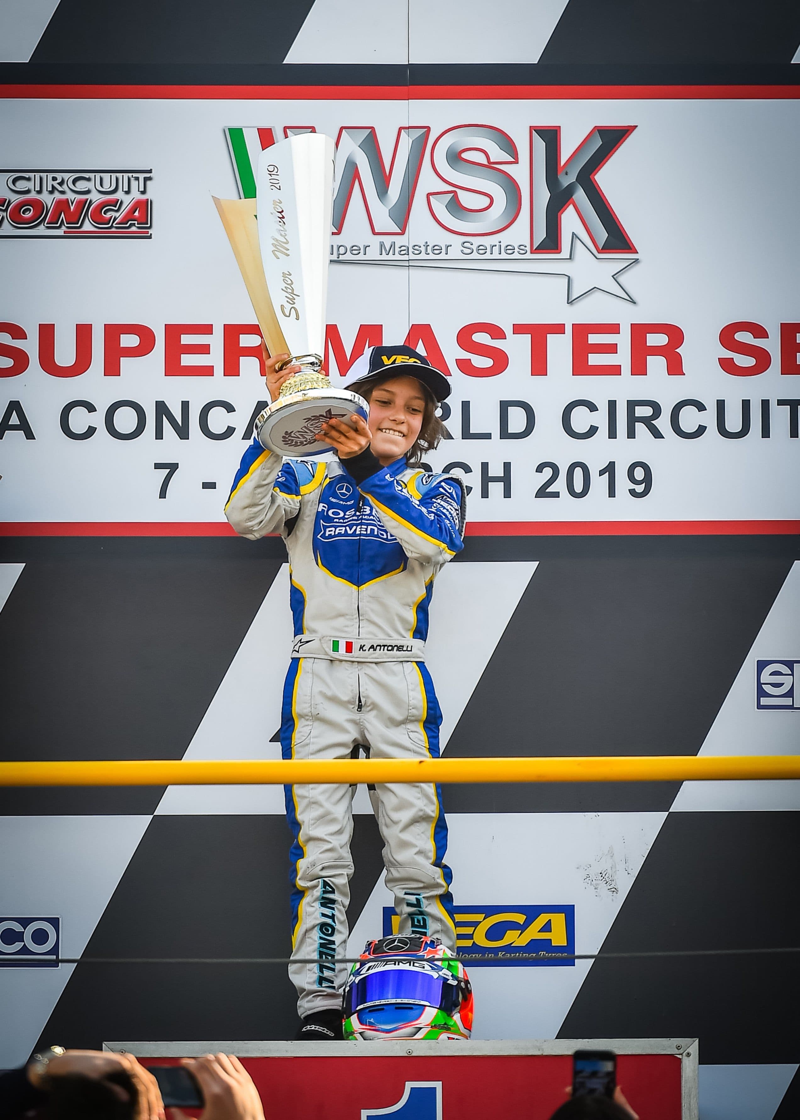 Young racer in blue and white suit joyfully holds large trophy on podium. Helmet with Italian flag colors rests below.