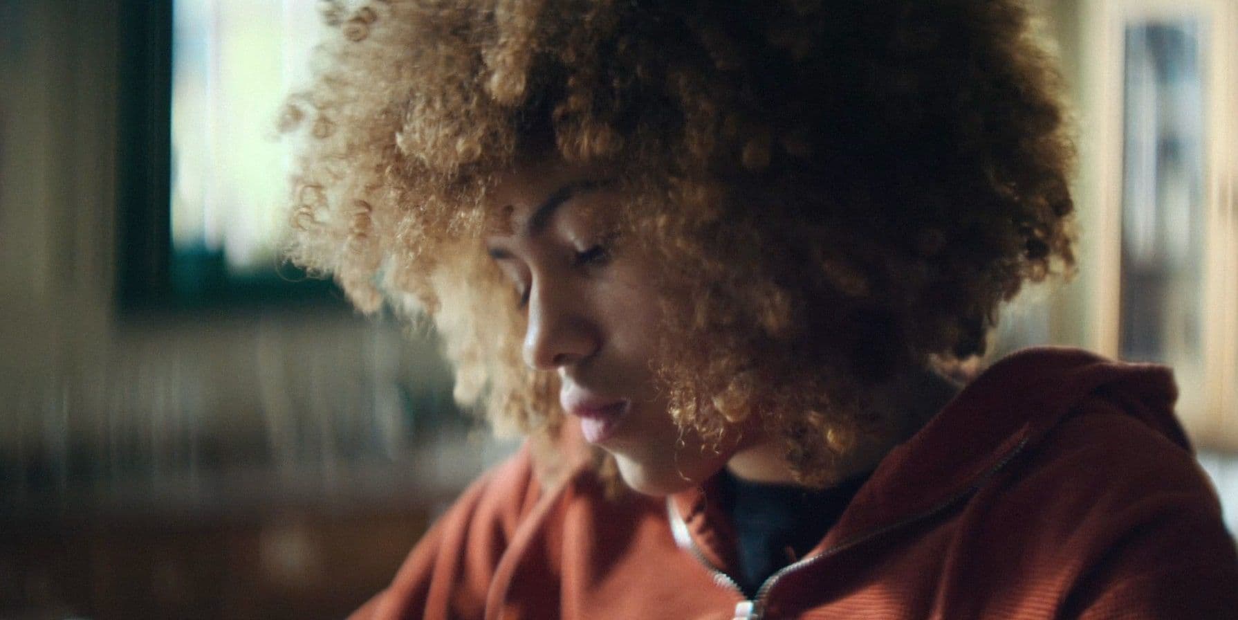 Young person with curly hair in a red hoodie, focused and thoughtful, indoors with blurred background, soft natural light.
