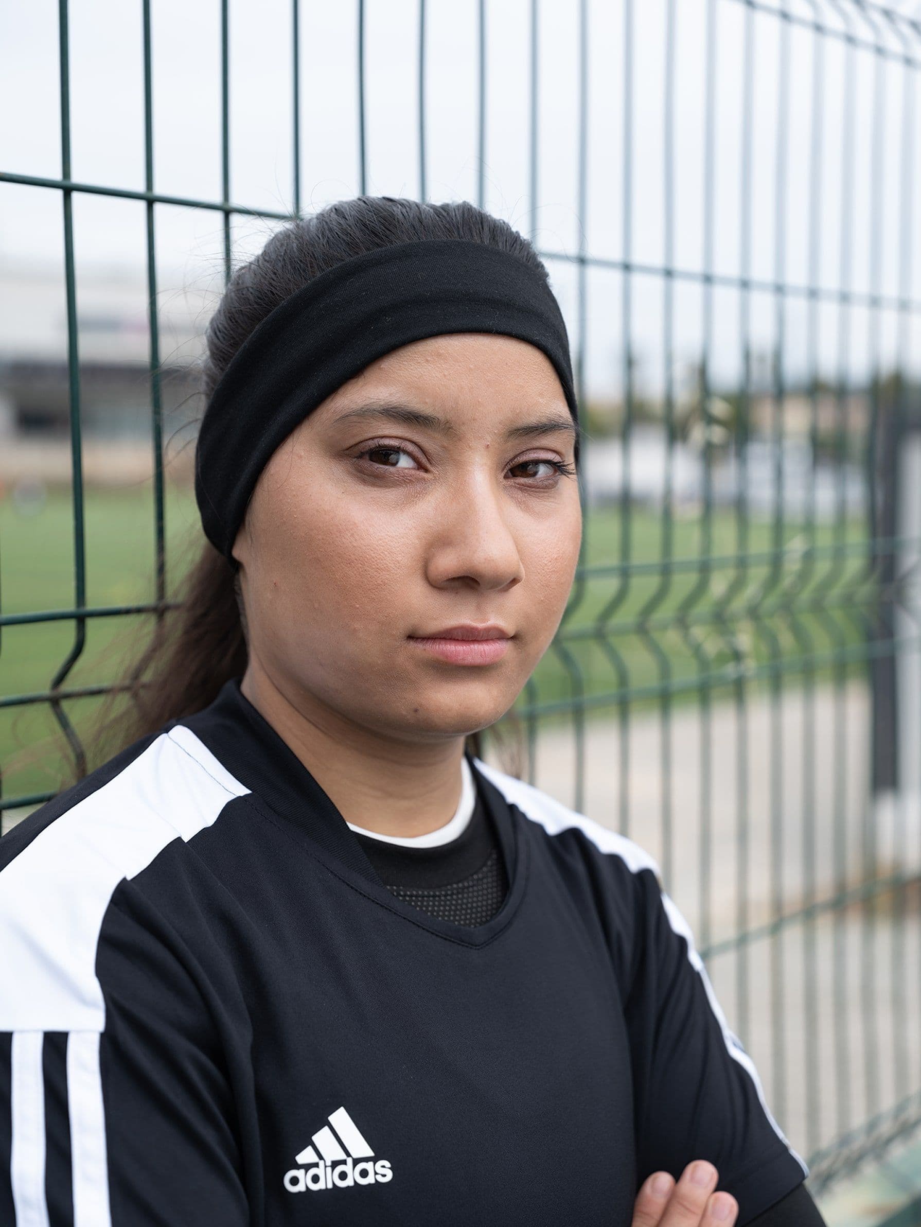 Young woman in black Adidas sports jersey and headband stands confidently by a wire fence on a soccer field.