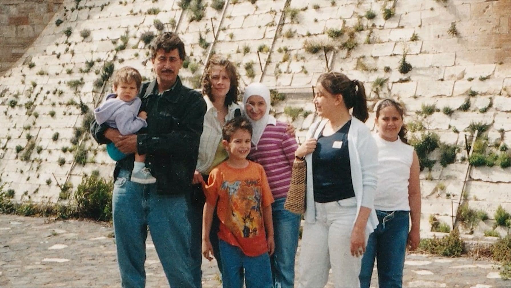 An old family photo of a group of seven people, including children and adults, stand smiling in front of a stone wall with greenery. Casual attire.