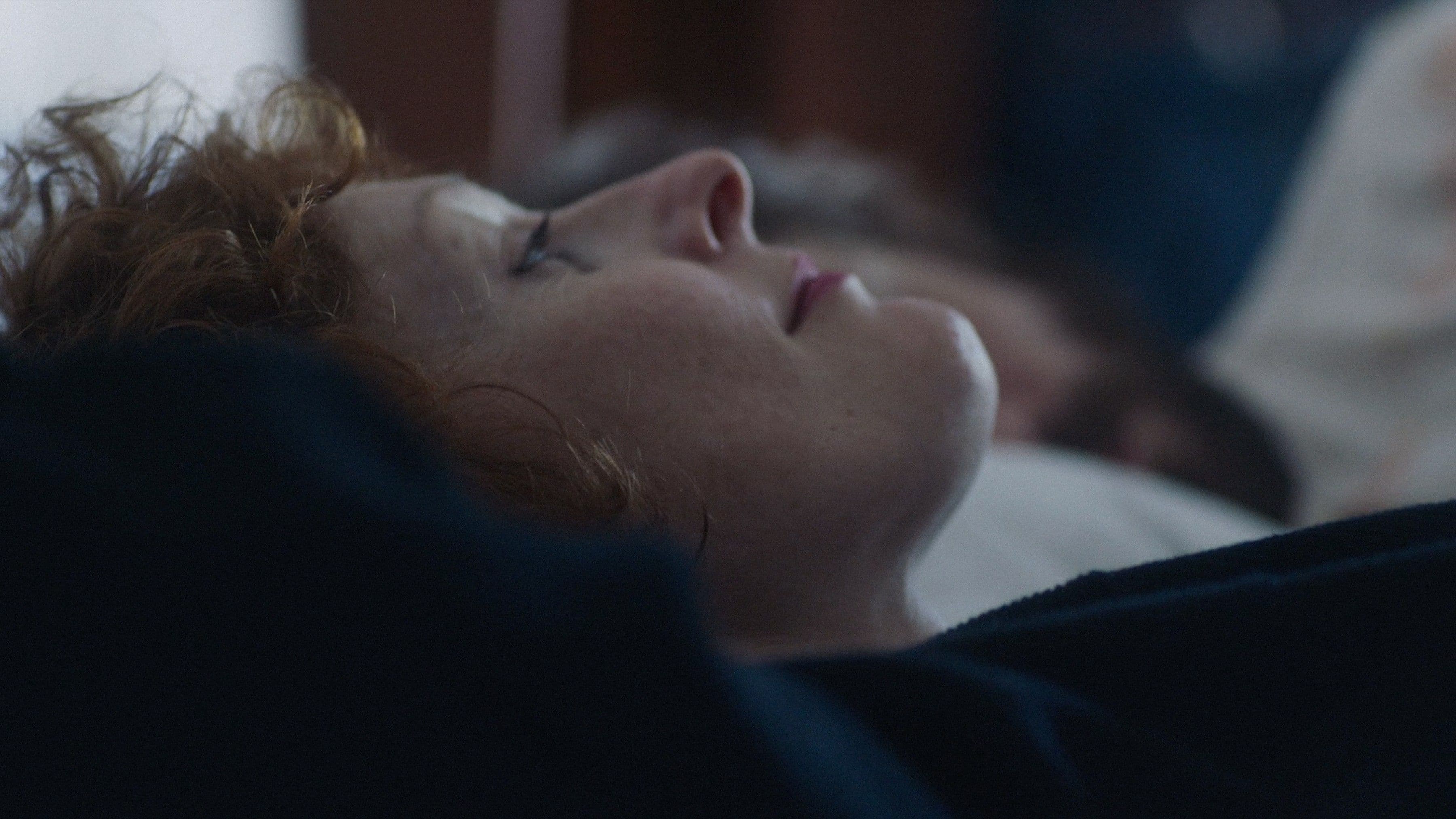 A woman with curly red hair lies on her back in bed, eyes closed, wearing a dark top. The background is softly blurred.