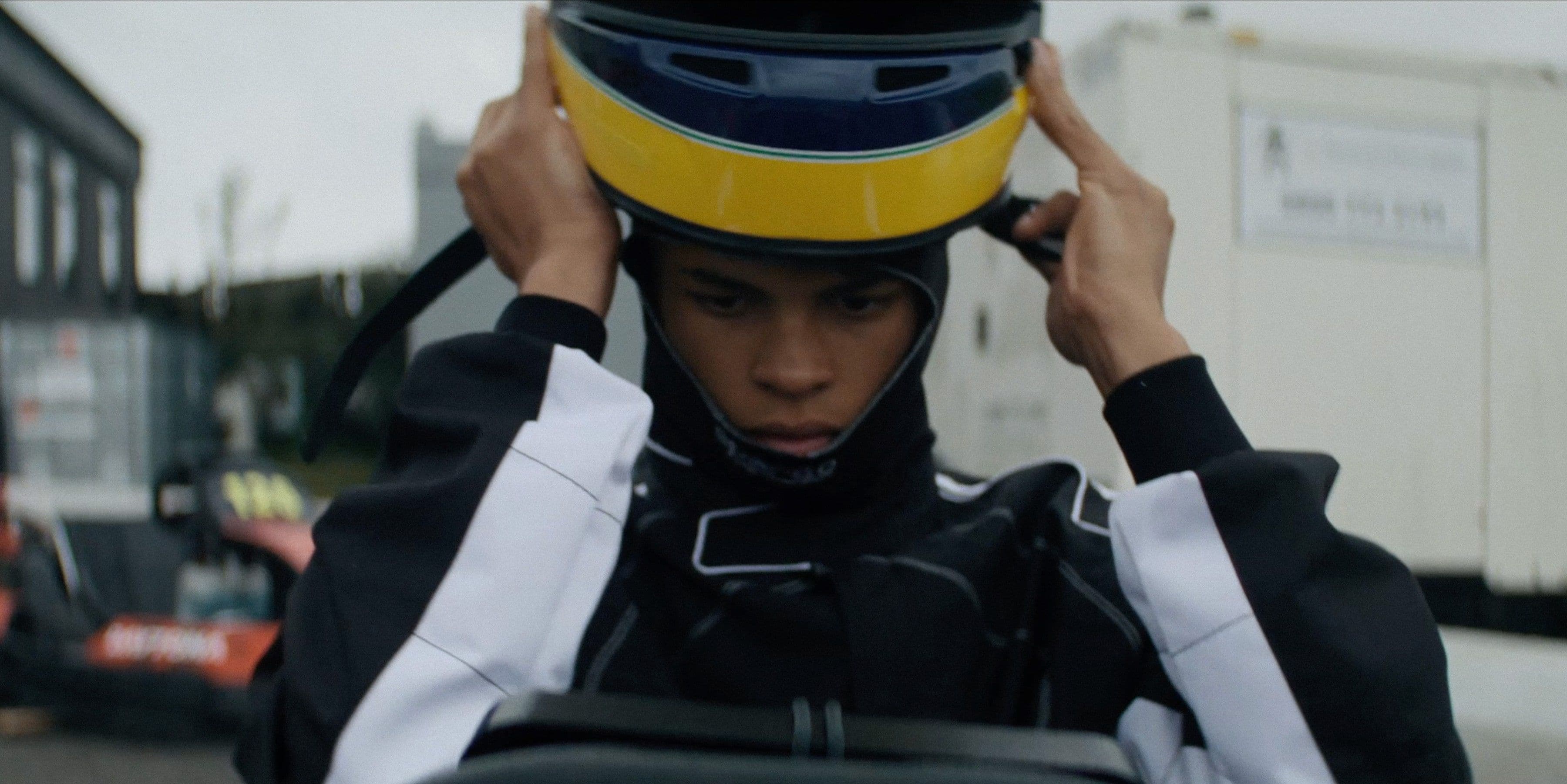 Person in black and white racing suit adjusts yellow and blue helmet. Putdoors near racing equipment and trailers.