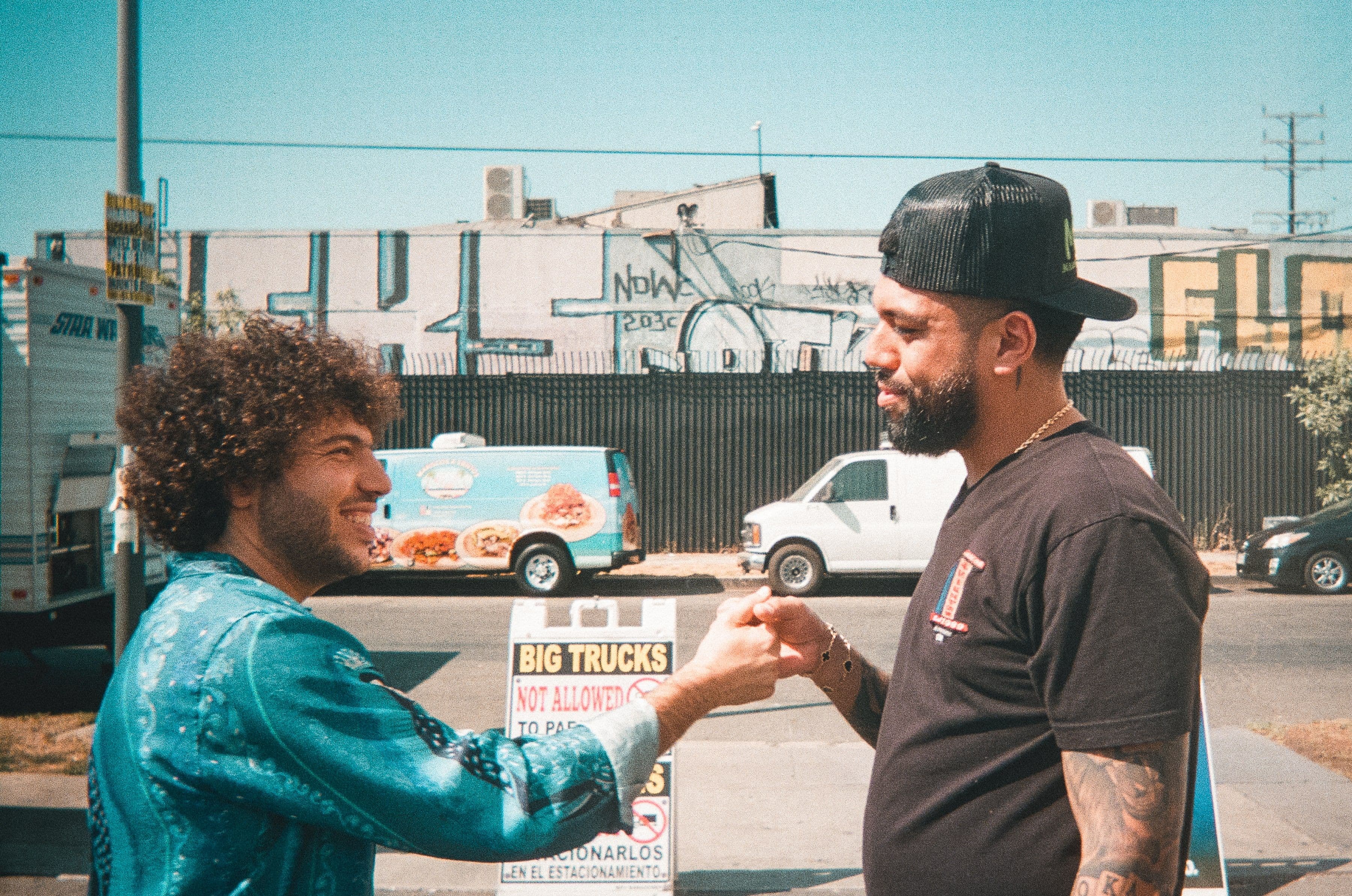 Two men fist bump on a sunny street. One wears a blue jacket, the other a black cap and shirt. Graffiti and vehicles in background.