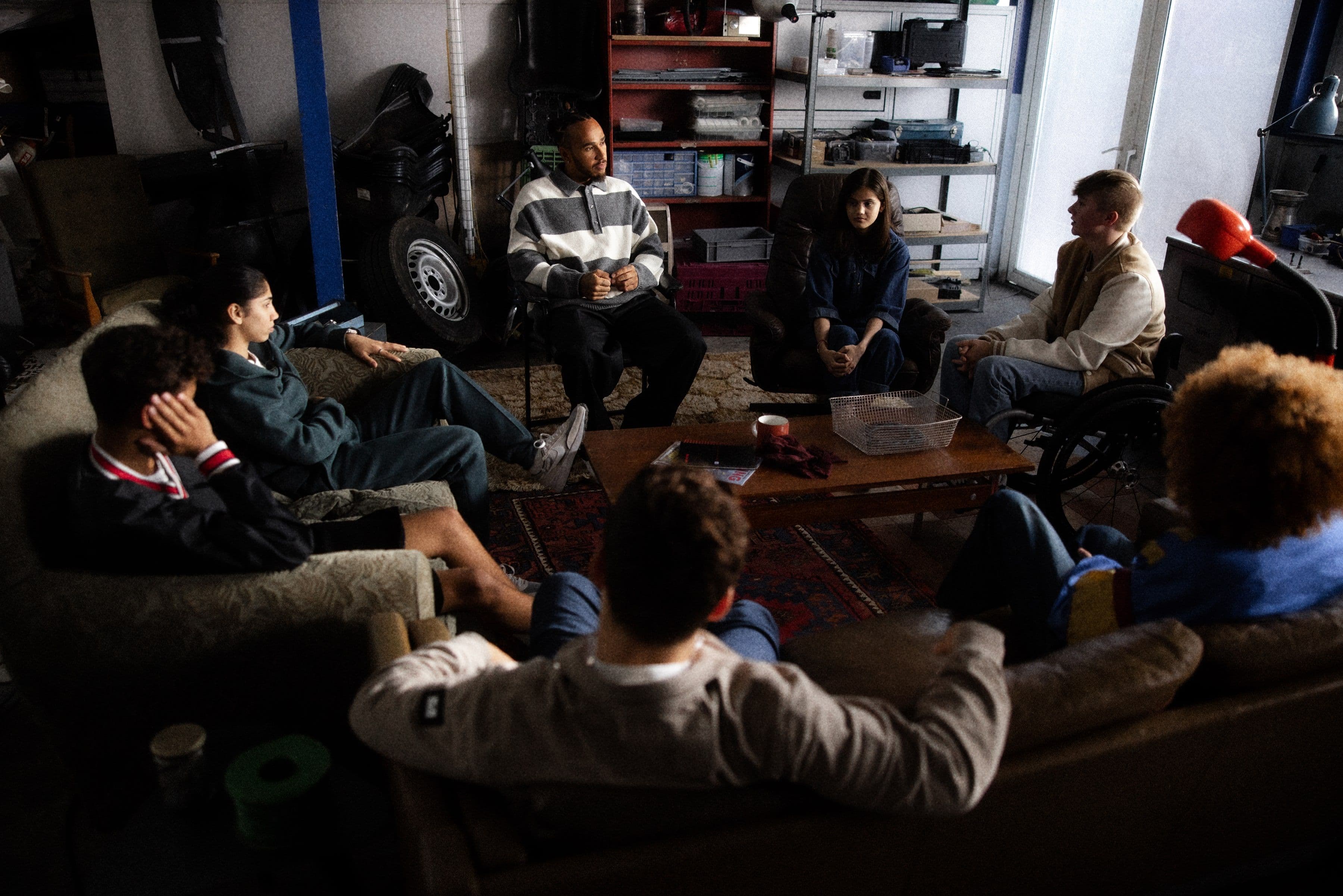 A diverse group of seven people sit in a circle in a dimly lit garage, engaged in conversation. Shelves and tools are in the background.