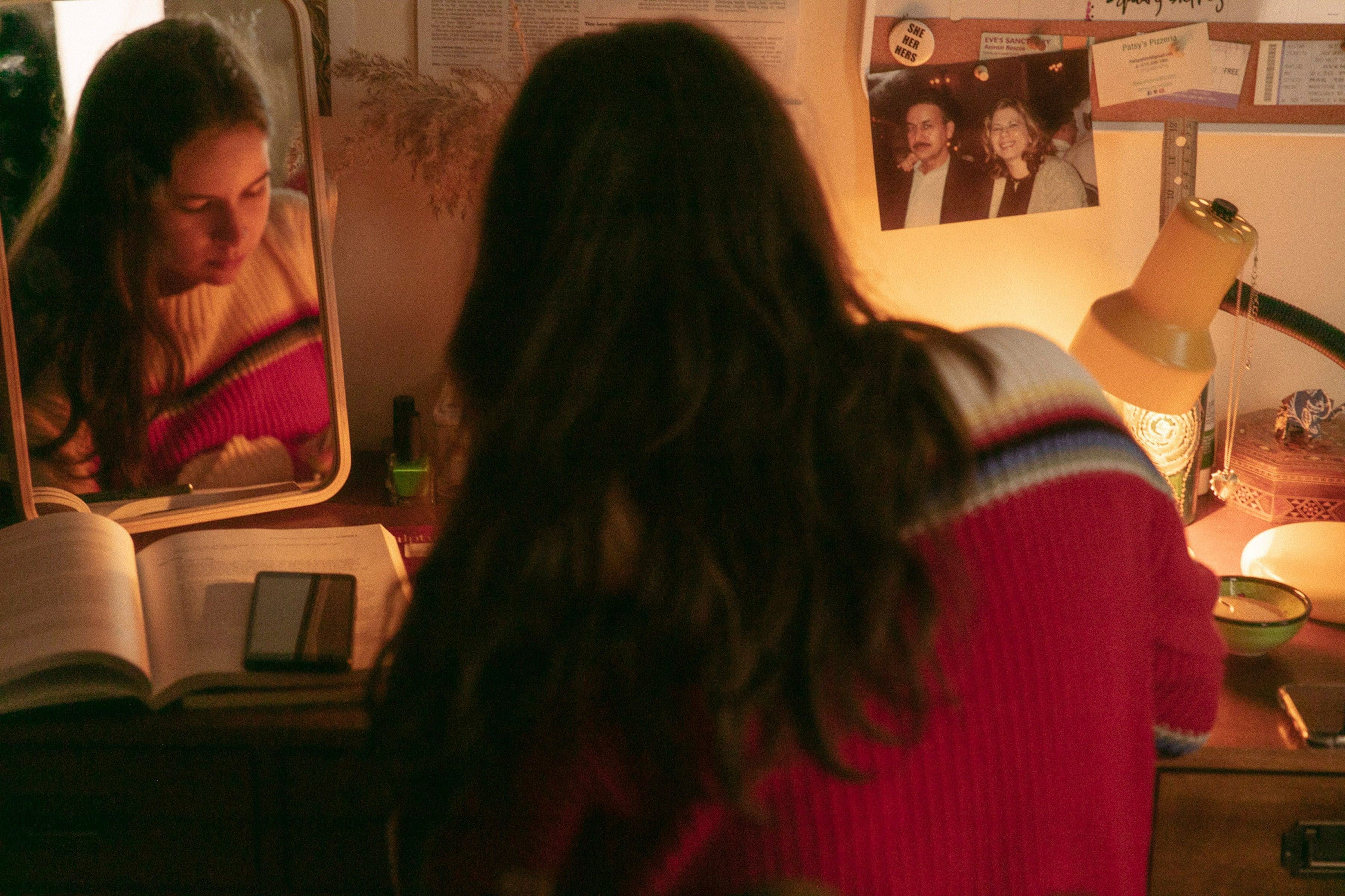 Woman in red sweater studies at a desk with an open book, phone, and lamp. Her reflection is visible in a mirror.