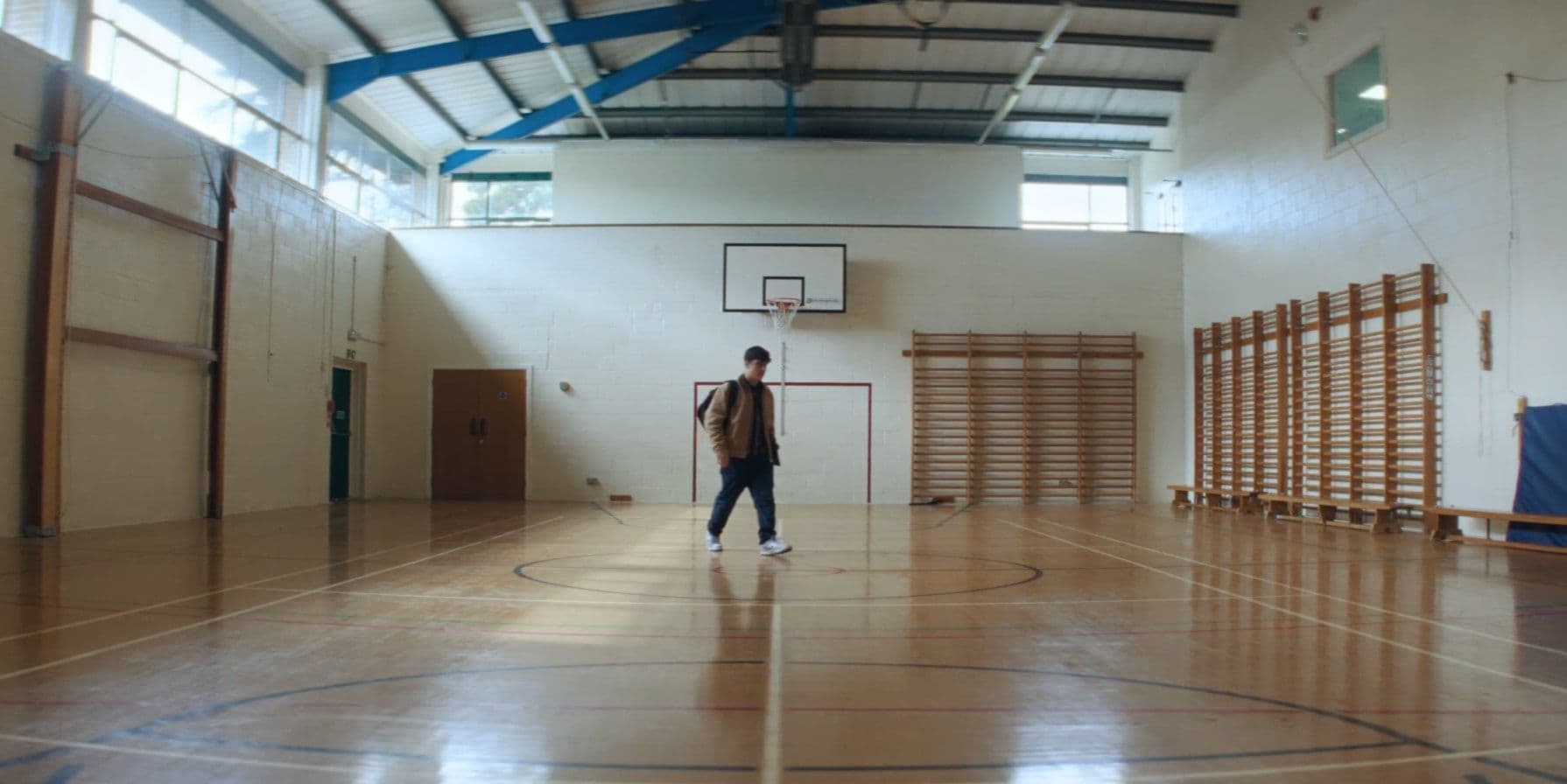 Man walking alone in an empty gymnasium with wooden floors, white walls, blue ceiling beams, and a basketball hoop.