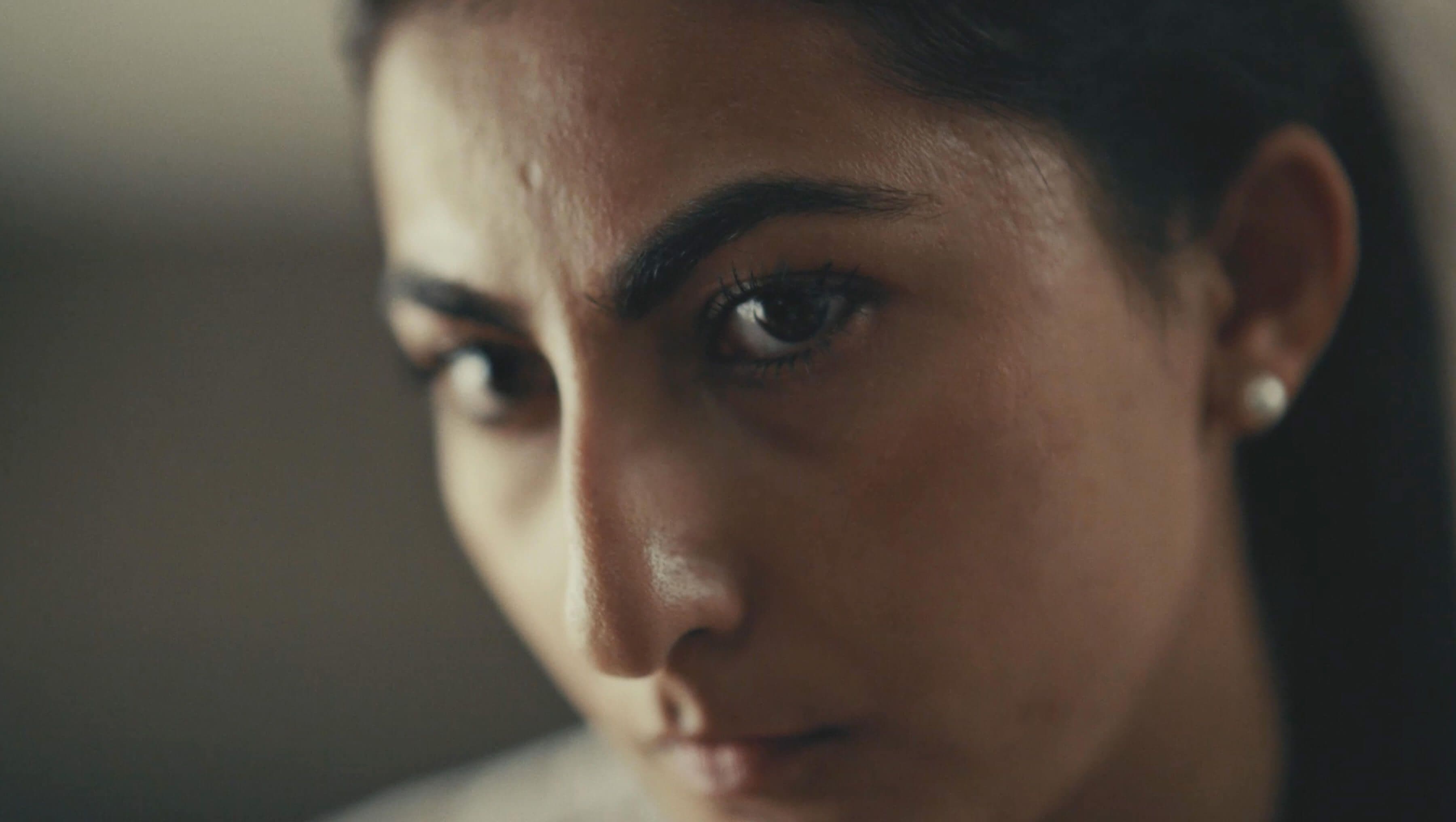 Close-up of a woman with intense expression, dark hair, and pearl earrings, in soft lighting, focusing on her eyes and face.