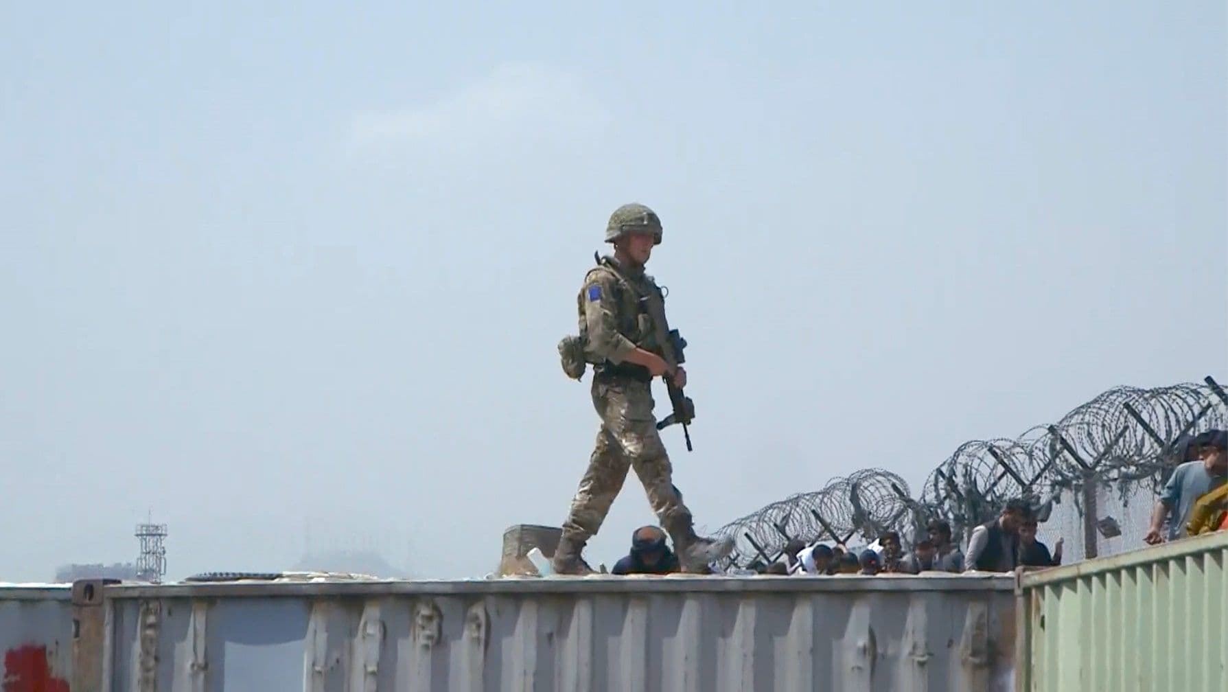 A soldier in camouflage and helmet walks on a shipping container, holding a rifle. Barbed wire and people are visible below.