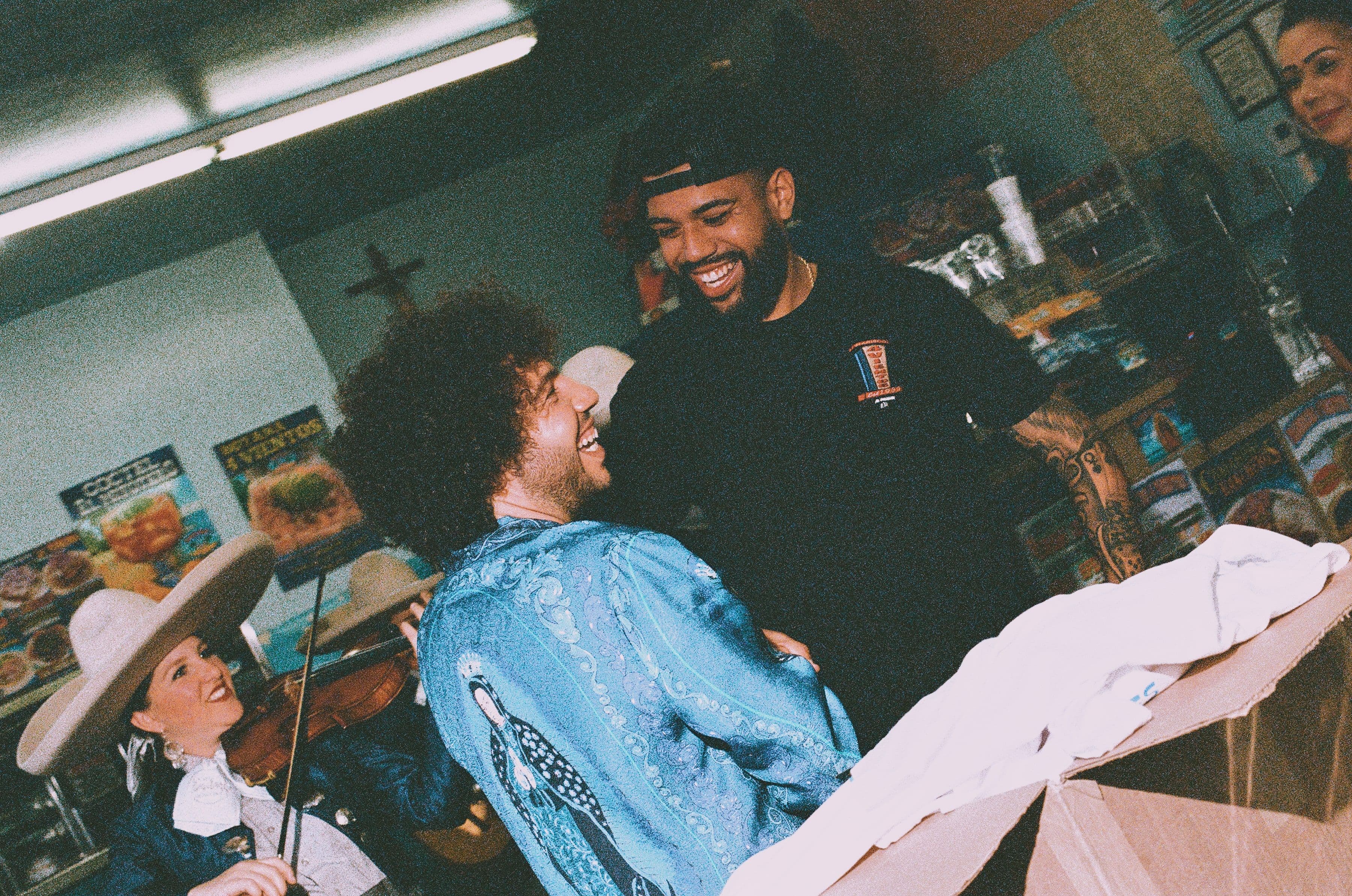 Two people laughing in a restaurant, one in a blue jacket, the other in a black shirt. A musician in a sombrero plays violin nearby.