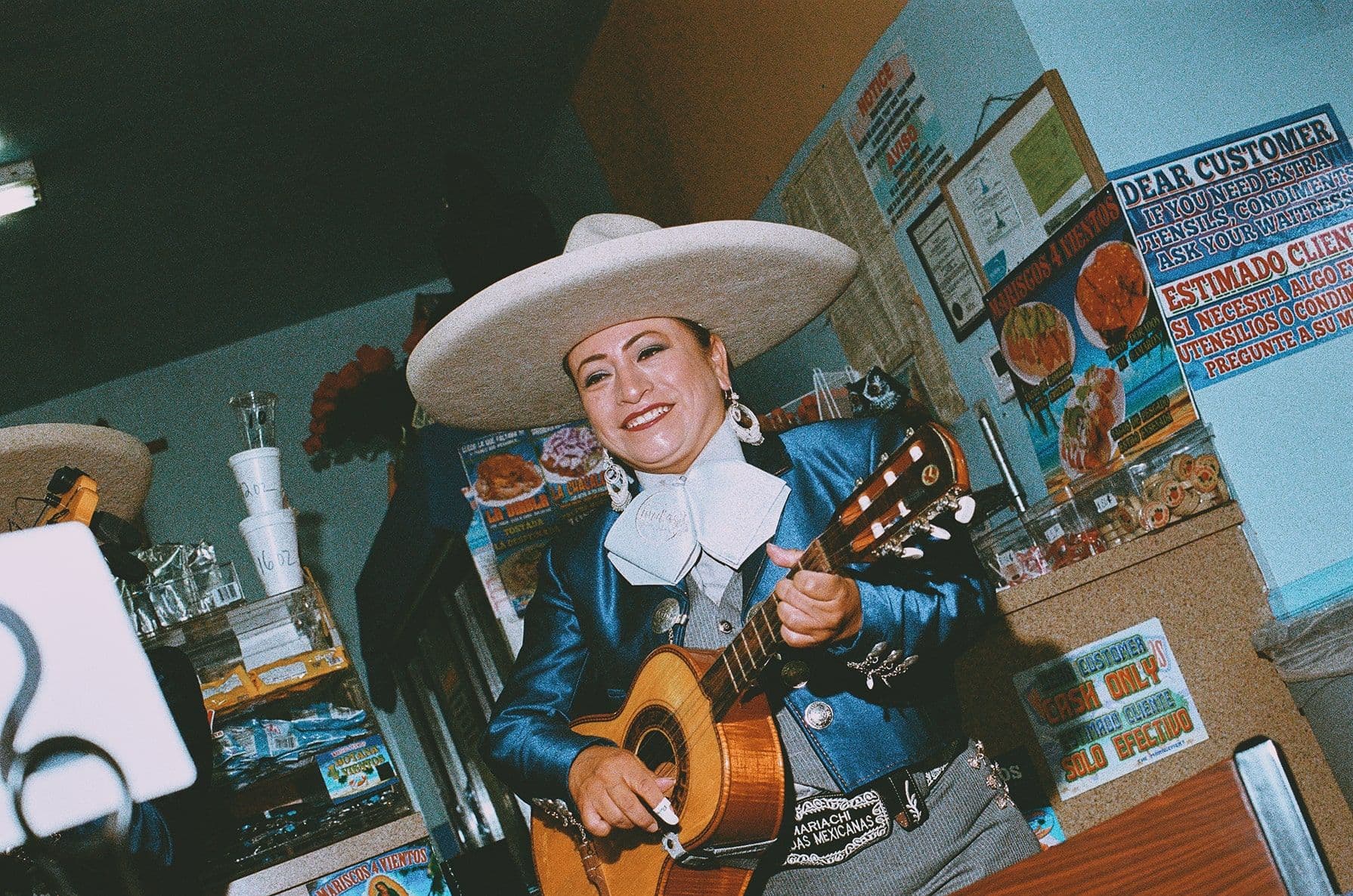 Smiling musician in blue mariachi outfit and white sombrero plays guitar in a colorful restaurant with posters and food items in the background.