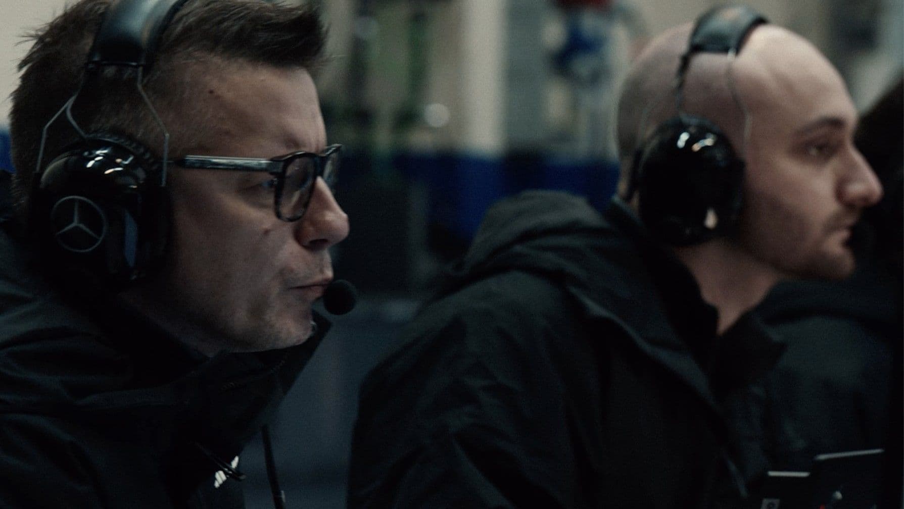 Two men wearing headsets and black jackets focus intently on racing results on screens. Garage setting.