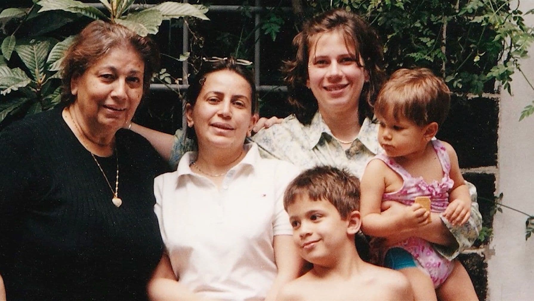 An old family photo of a group of five people, including three women and two young kids, stand together outdoors near green foliage.